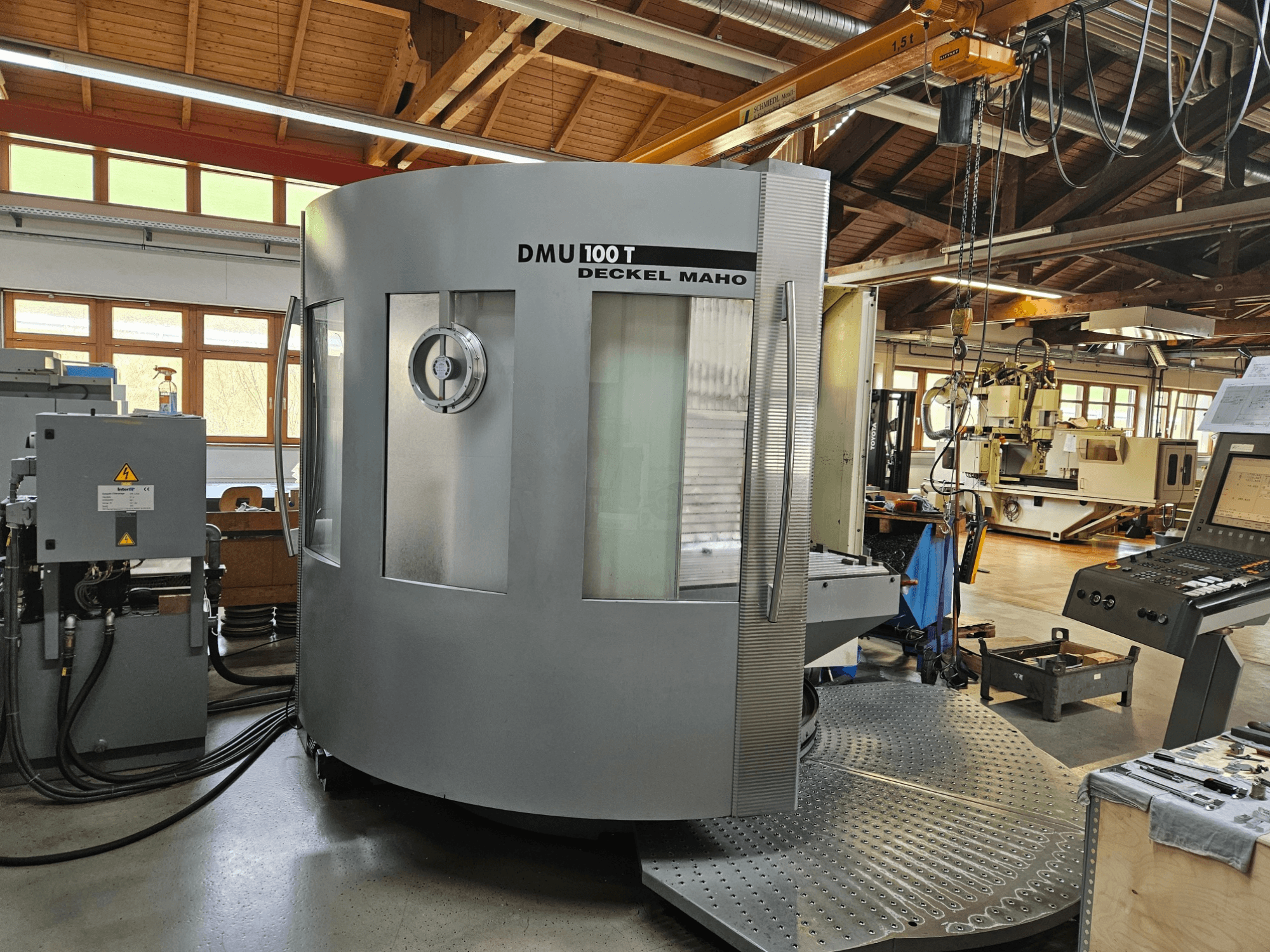DMU 100 T Deckel Maho CNC mašīna no priekšpuses, ar lielu apaļu pamatni, vadības paneli un stikla korpusu.