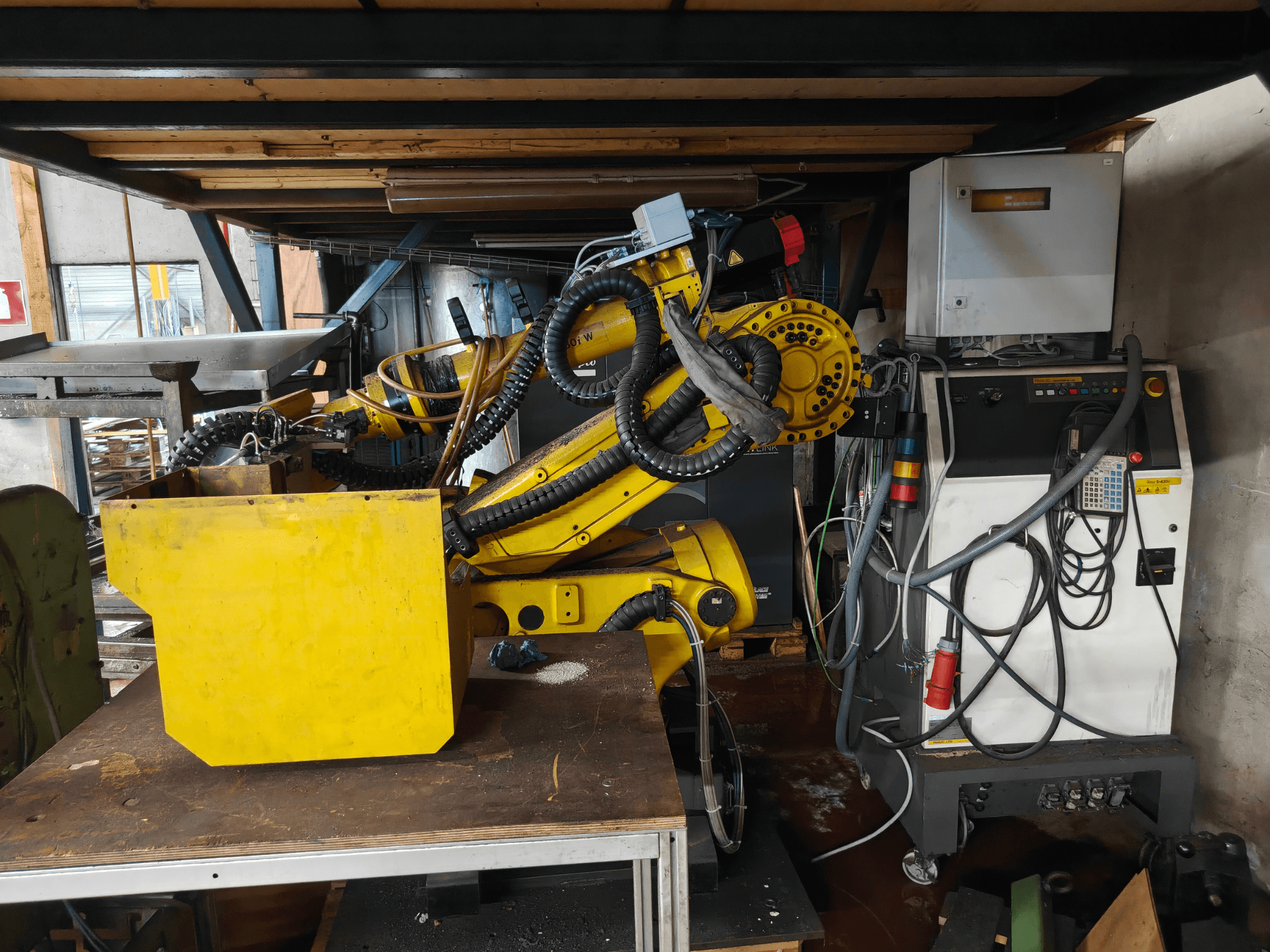 FANUC robota S-430i W rokas priekšskats — dzeltenā krāsā ar vadības paneli, kabeļiem un dzeltenu detaļu uz darbgalda.