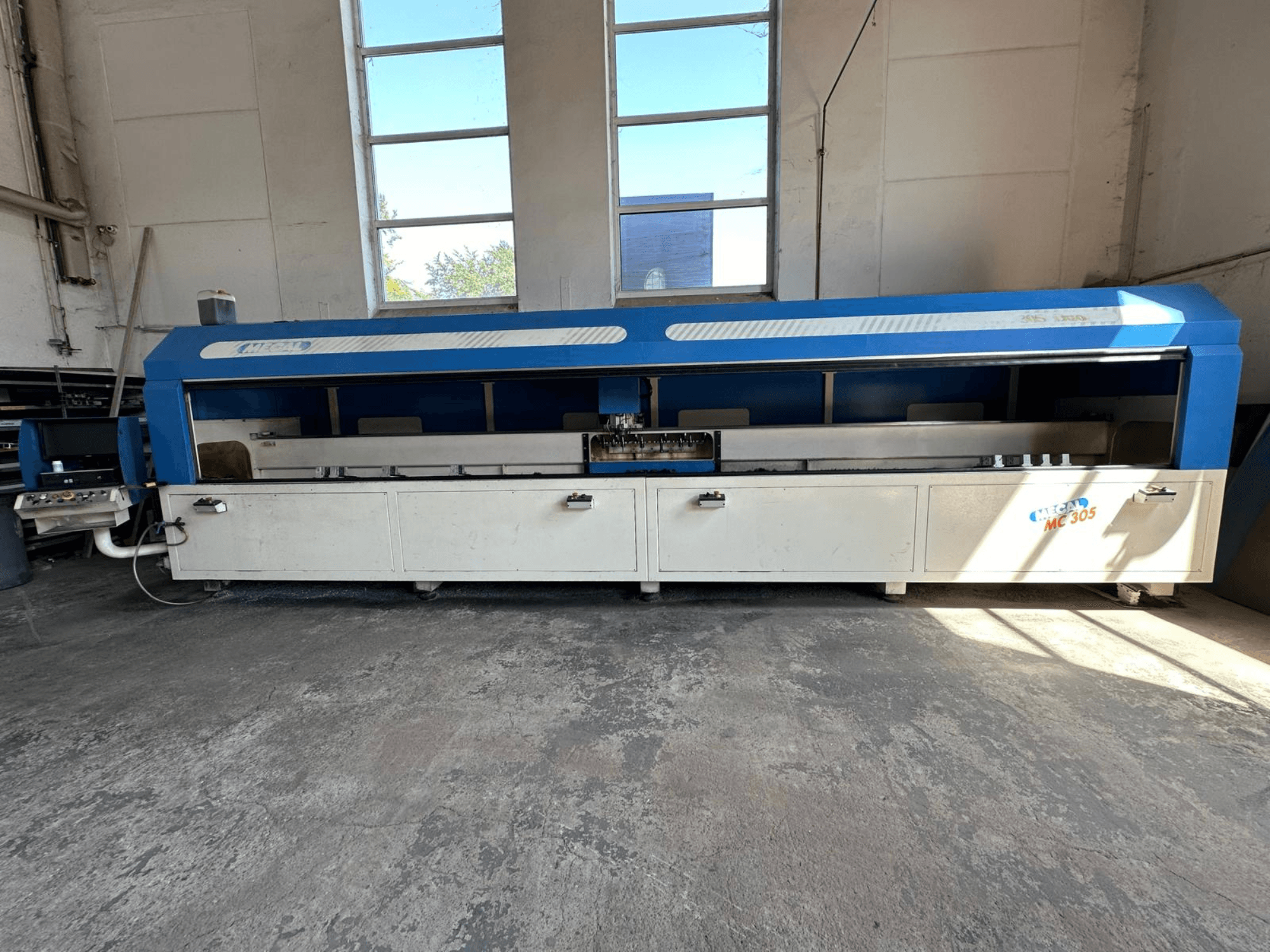 MECAL MC 305 vertikālā apstrādes centra priekšējais skats ar zili baltu dizainu, redzamu darba galdu un vadības paneli.