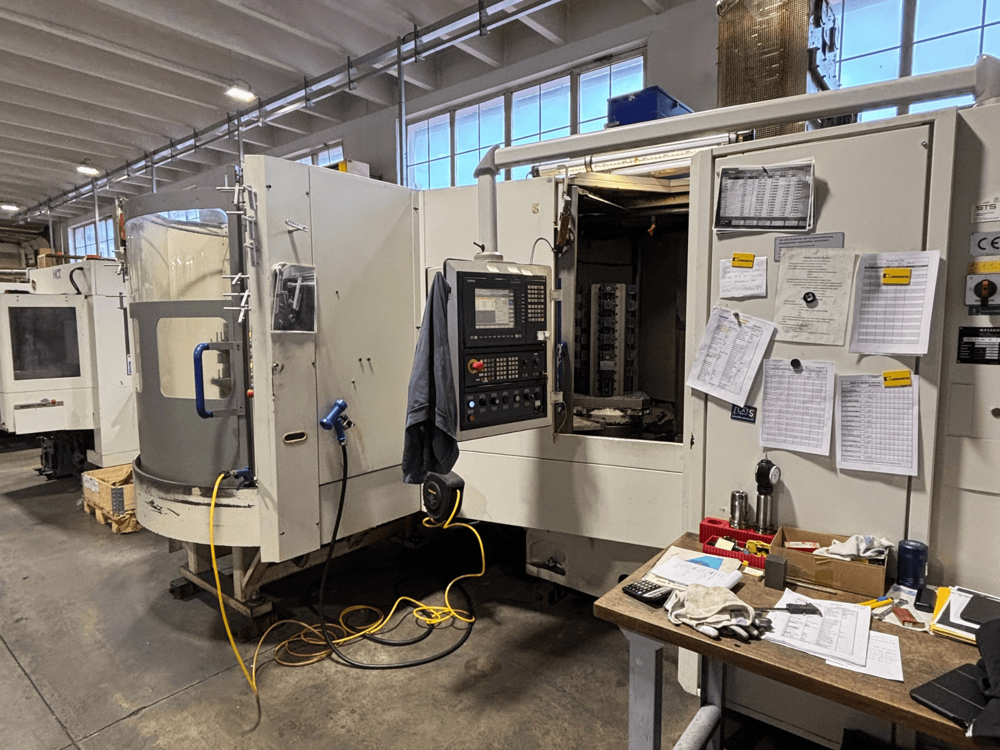 CNC darbnīca darbnīcā, skats no priekšpuses, vadības panelis un dokumentācija sānos; instrumenti un materiāli uz galda.
