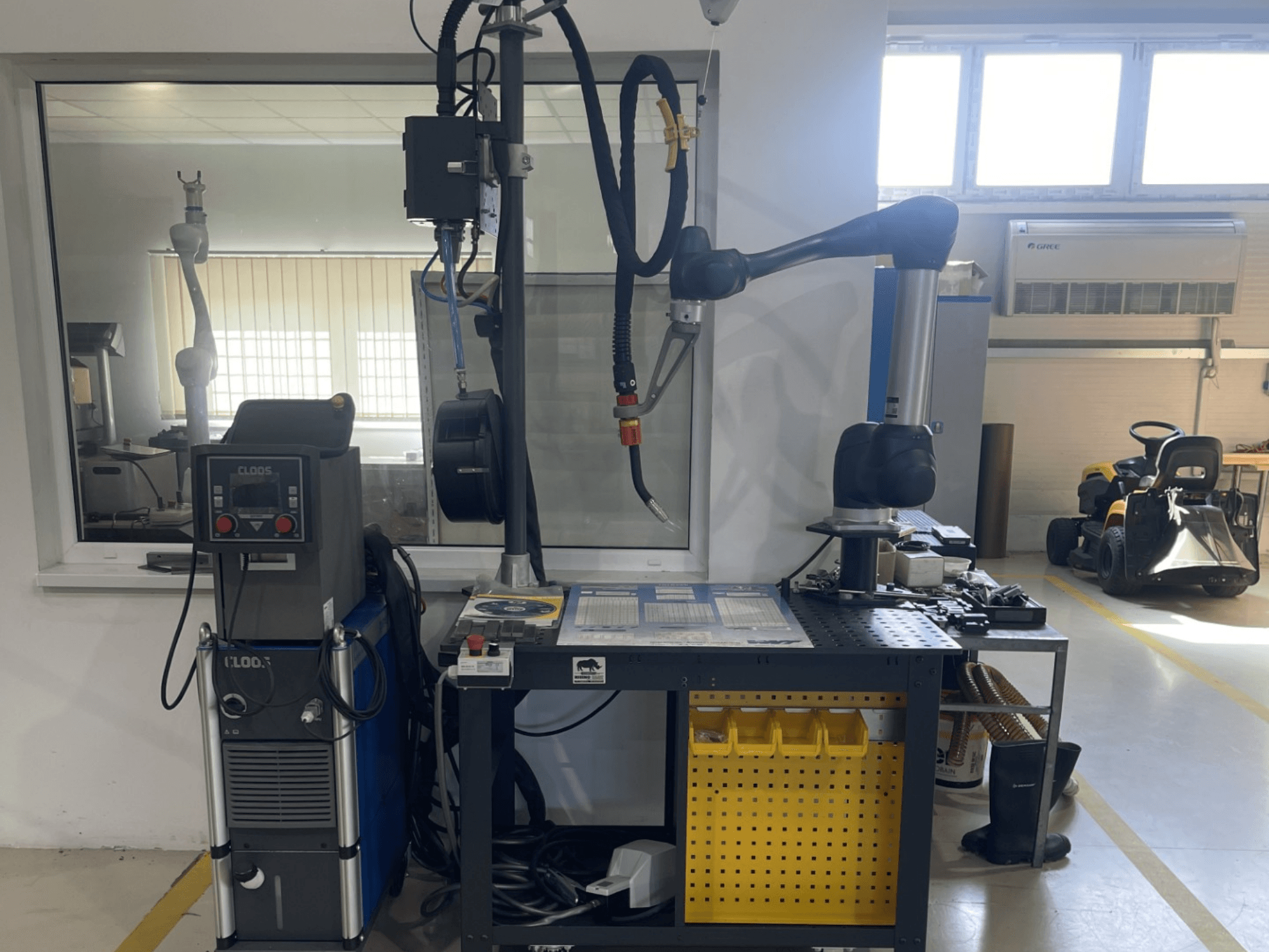 DOOSAN M1013 sadarbības robots darbnīcā, sānskats ar metināšanas iekārtām un instrumentiem uz darba galda.