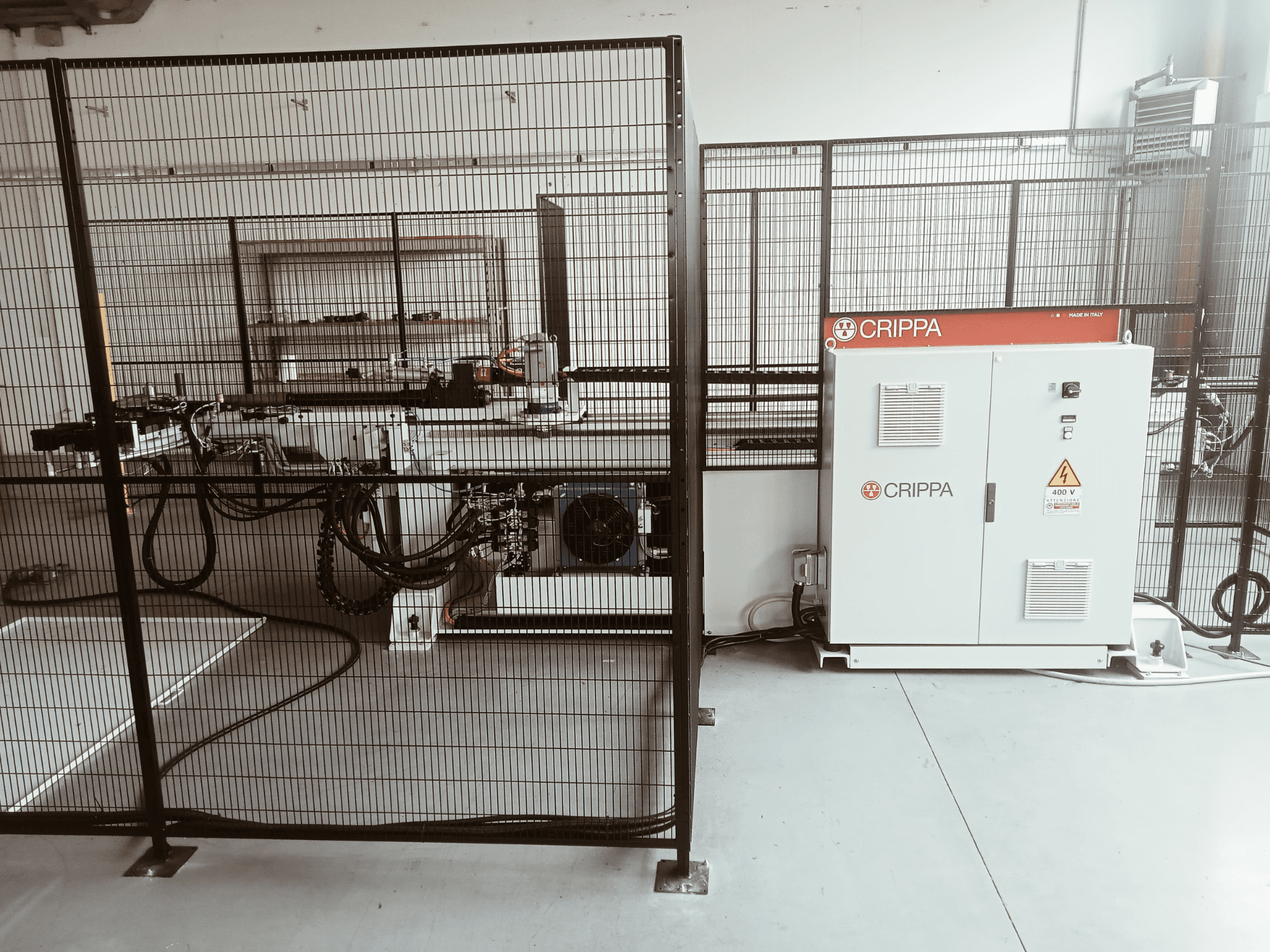 Balta CRIPPA mašīna aiz drošības būra, ar redzamām šļūtenēm un detaļām, skats no sāniem, industriālā vidē.