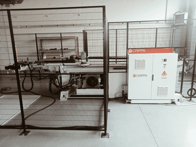 Balta CRIPPA mašīna aiz drošības būra, ar redzamām šļūtenēm un detaļām, skats no sāniem, industriālā vidē.