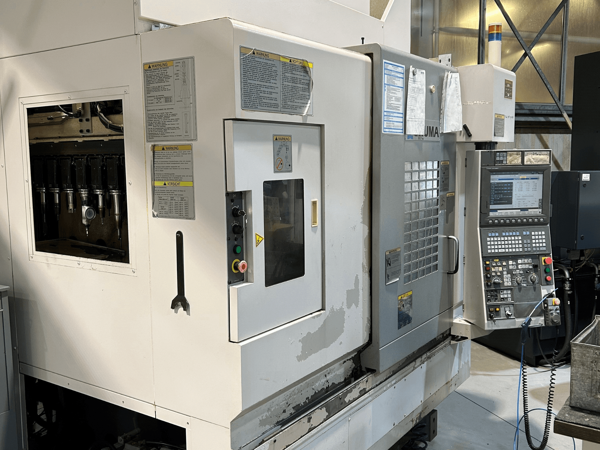 UMA CNC mašīnas priekšējais skats ar redzamiem instrumentu turētājiem, vadības paneli, brīdinājuma uzrakstiem un vadības pogām.
