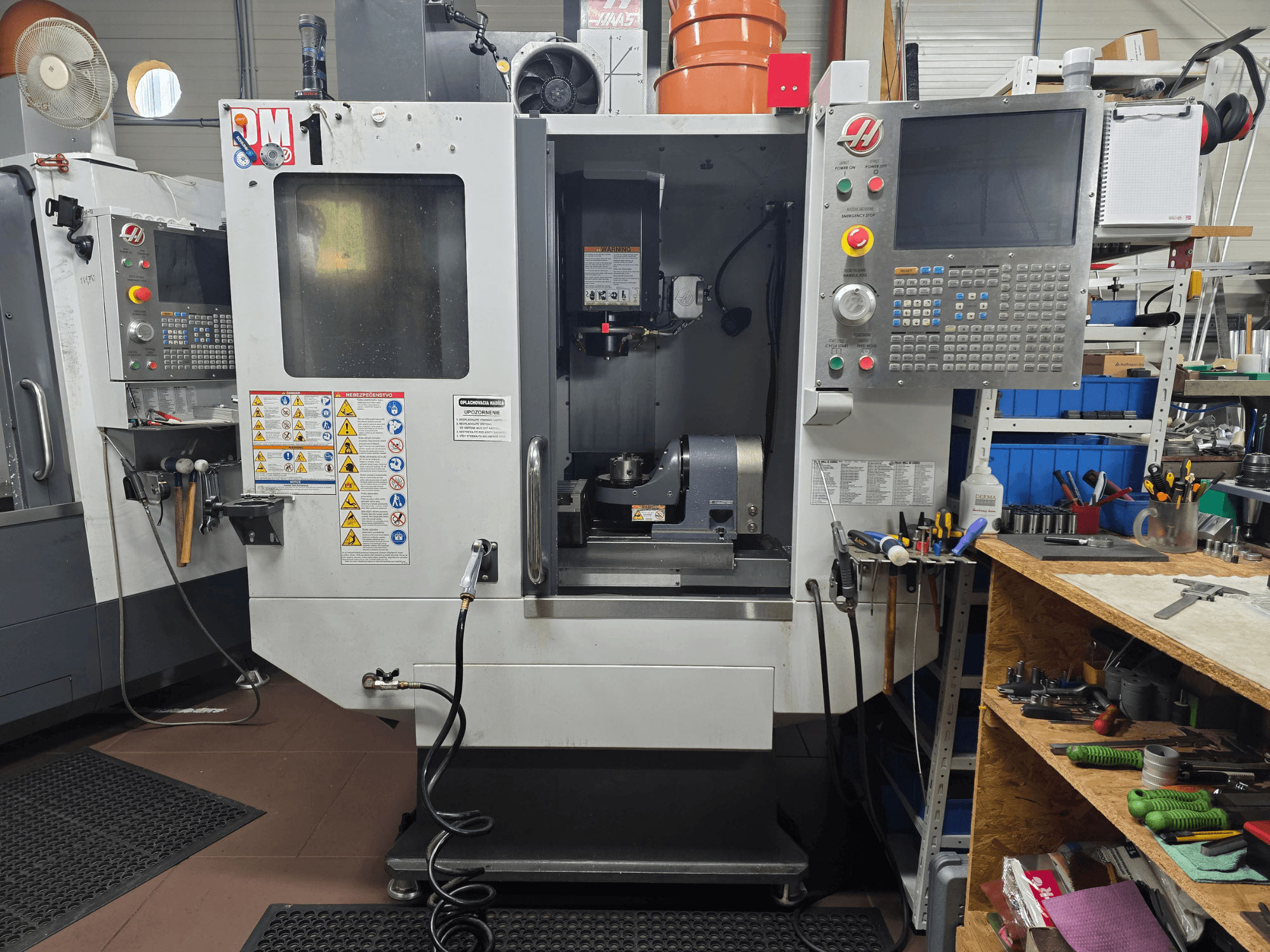Haas CNC darbgalds, modelis VF-2, skats no priekšpuses, kurā redzamas vadības ierīces, vārpstas zona, drošības marķējumi un instrumenti uz blakus esošā darbgalda.