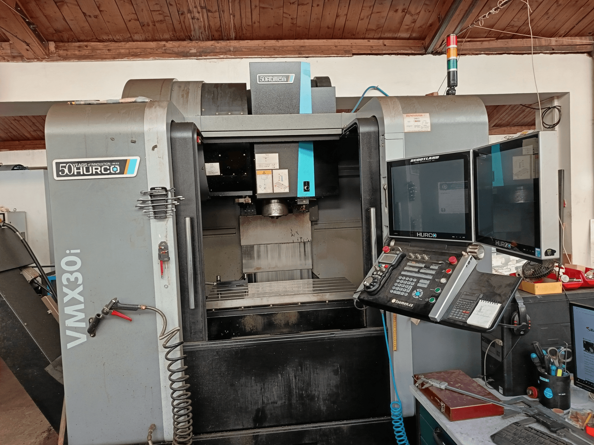 Hurco VMX30i CNC frēzmašīna priekšpusē ar diviem monitoriem, vadības paneli un pievienotu šļūteni.