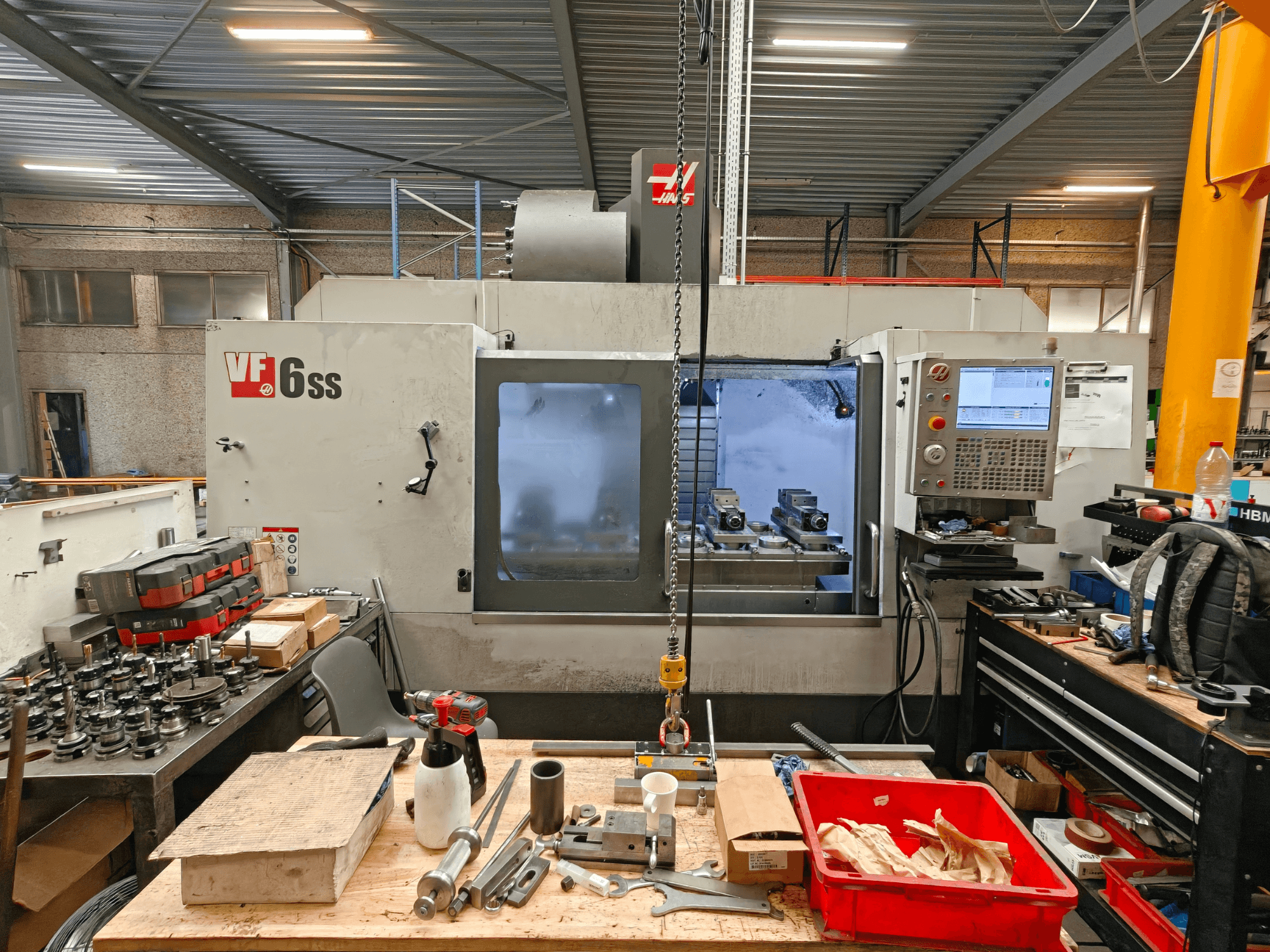 Vertikālais apstrādes centrs „Haas VF-6SS“ darbnīcā, skats no sāniem, kurā redzams vadības panelis un darba virsma, kas pārklāta ar instrumentiem.