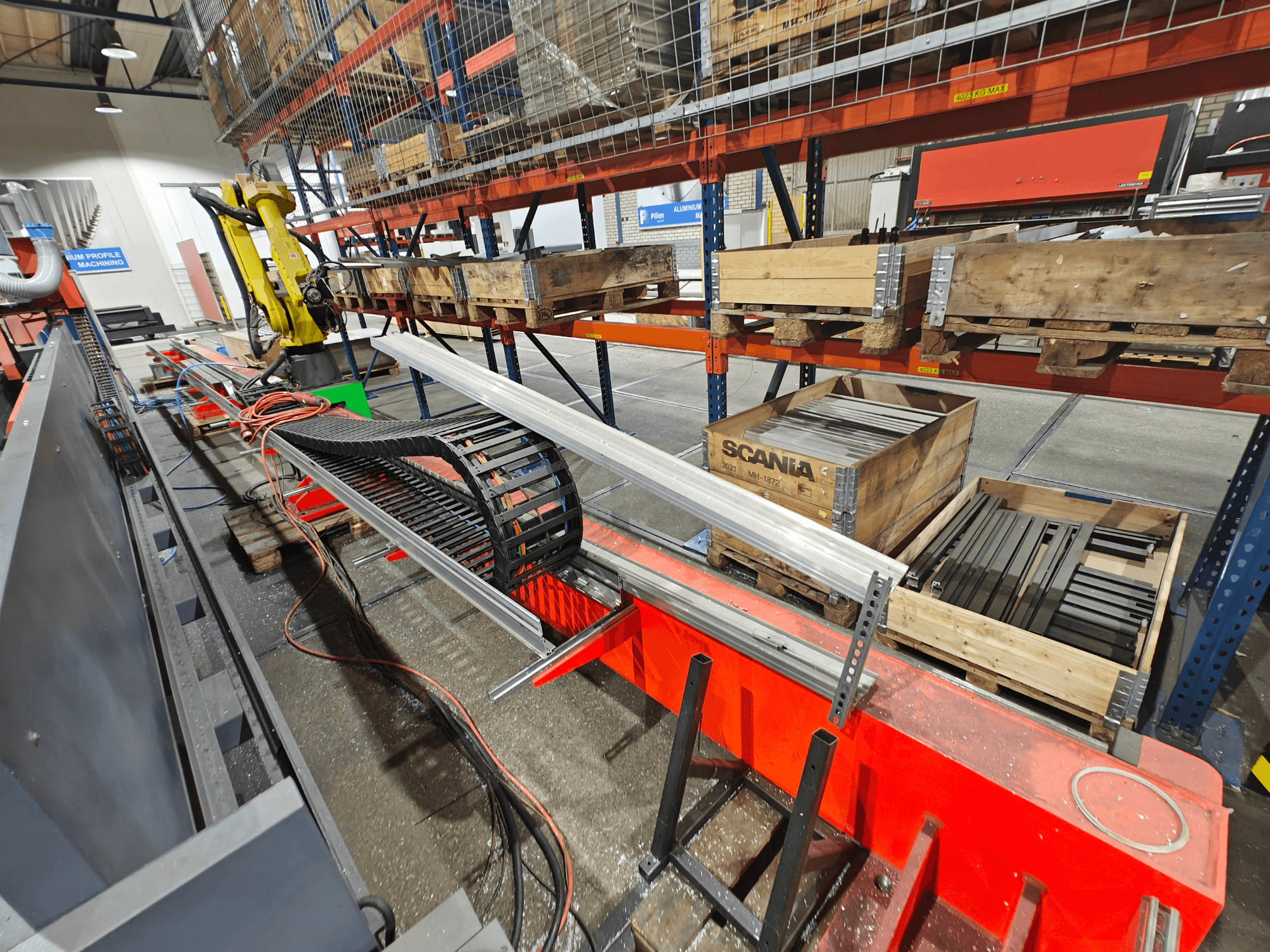 Robotizēta roka uz darbstacijas, redzami oranži un melni kabeļi, ar Scania kastēm un alumīnija profiliem rūpnīcā.