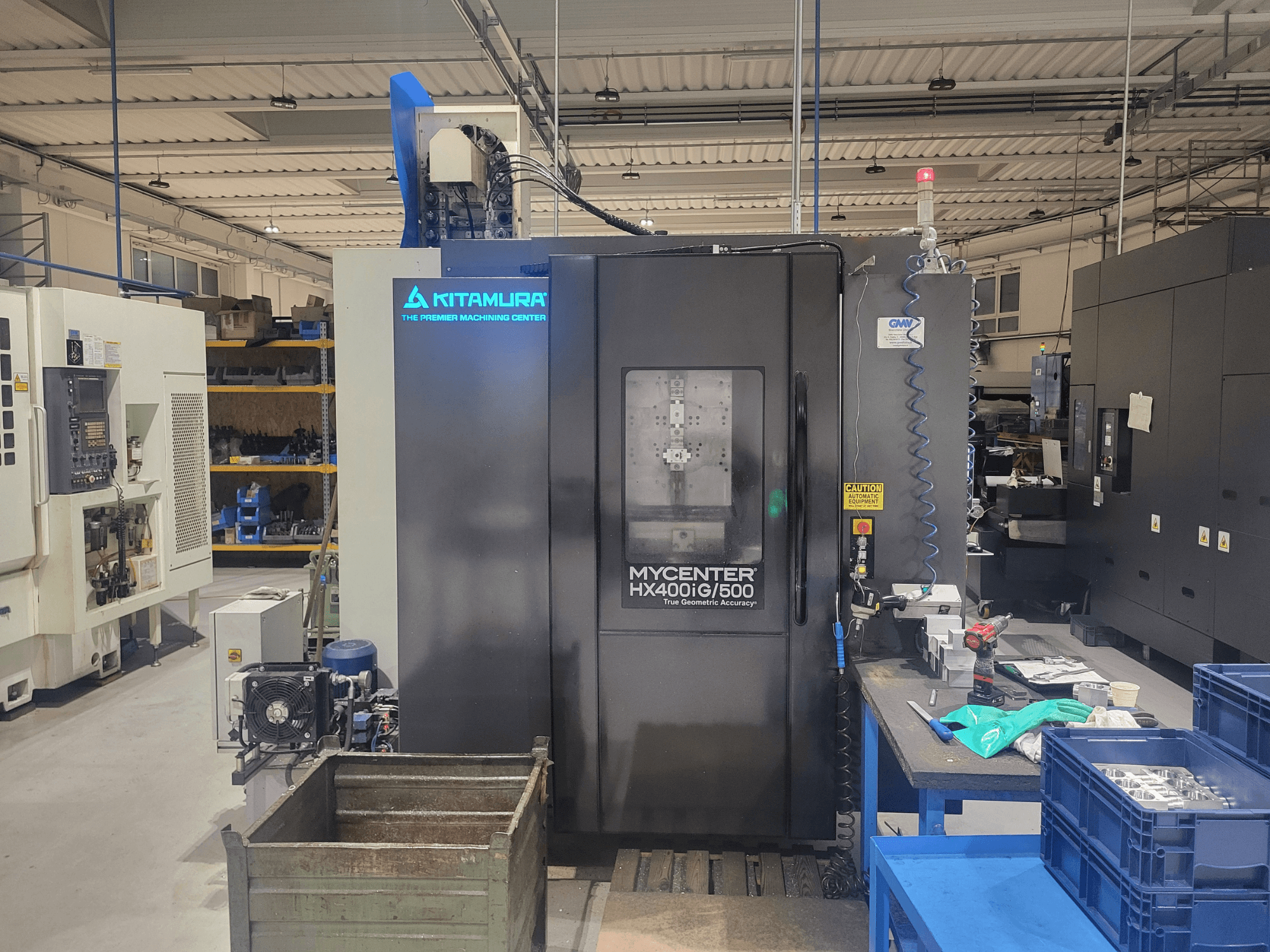 Kitamura Mycenter HX400iG/500 CNC mašīna darbnīcā, skats no priekšpuses, ar vadības ierīcēm, instrumentu noliktavu un materiāliem blakus.