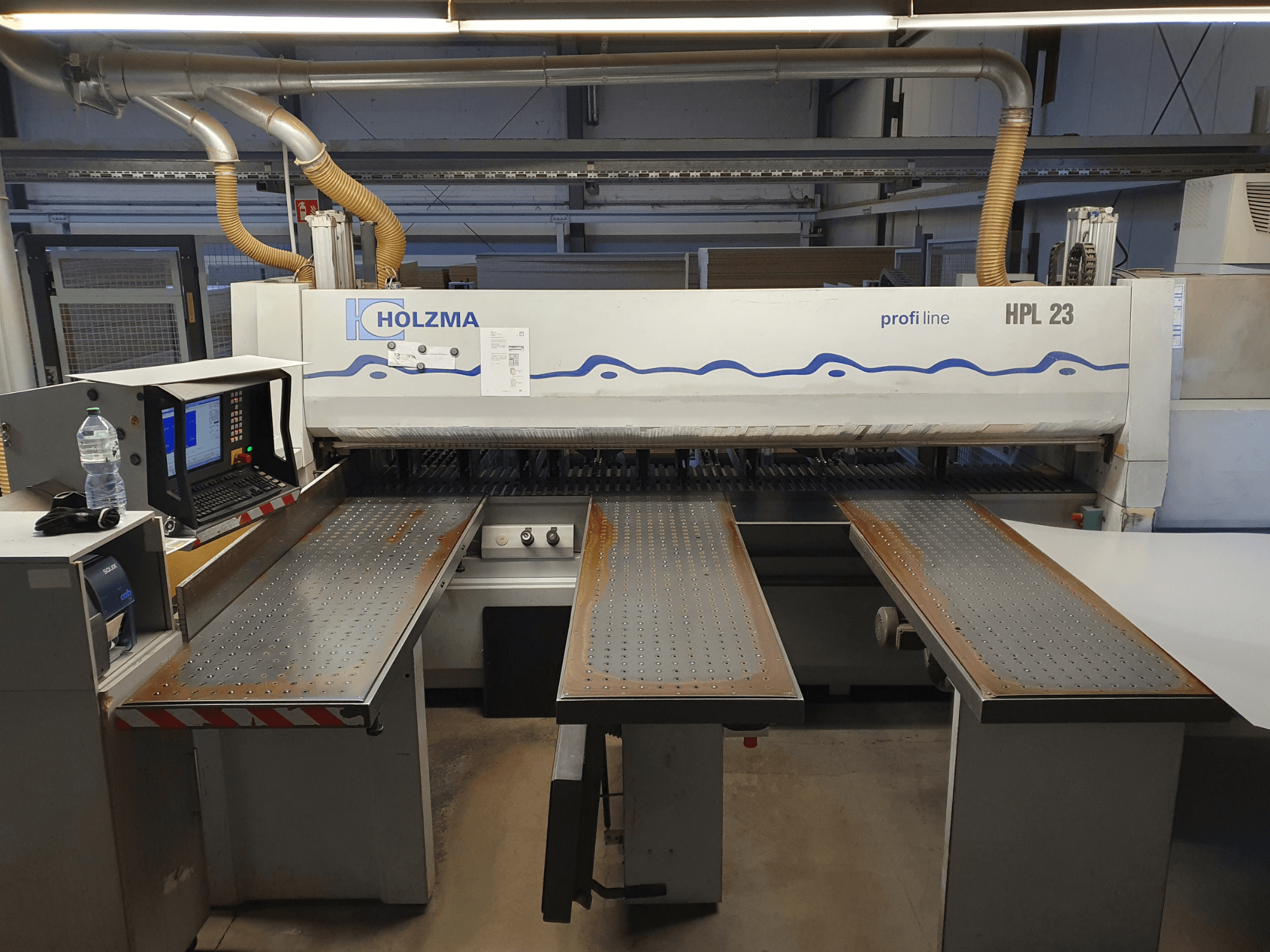 HOLZMA HPL 23 griešanas iekārta no priekšpuses ar trim perforētiem galdiem, datora termināli un ūdens pudeli blakus.