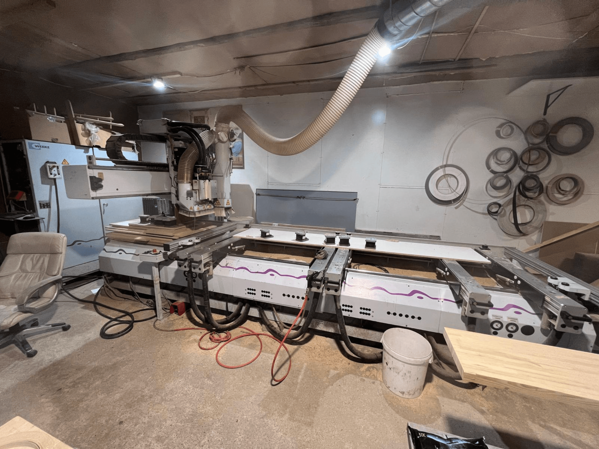 CNC kokapstrādes centrs „Weeke OPTIMAT BP 120” darbnīcā, skats no priekšpuses, uz virsmas redzami koka gabali un instrumenti.