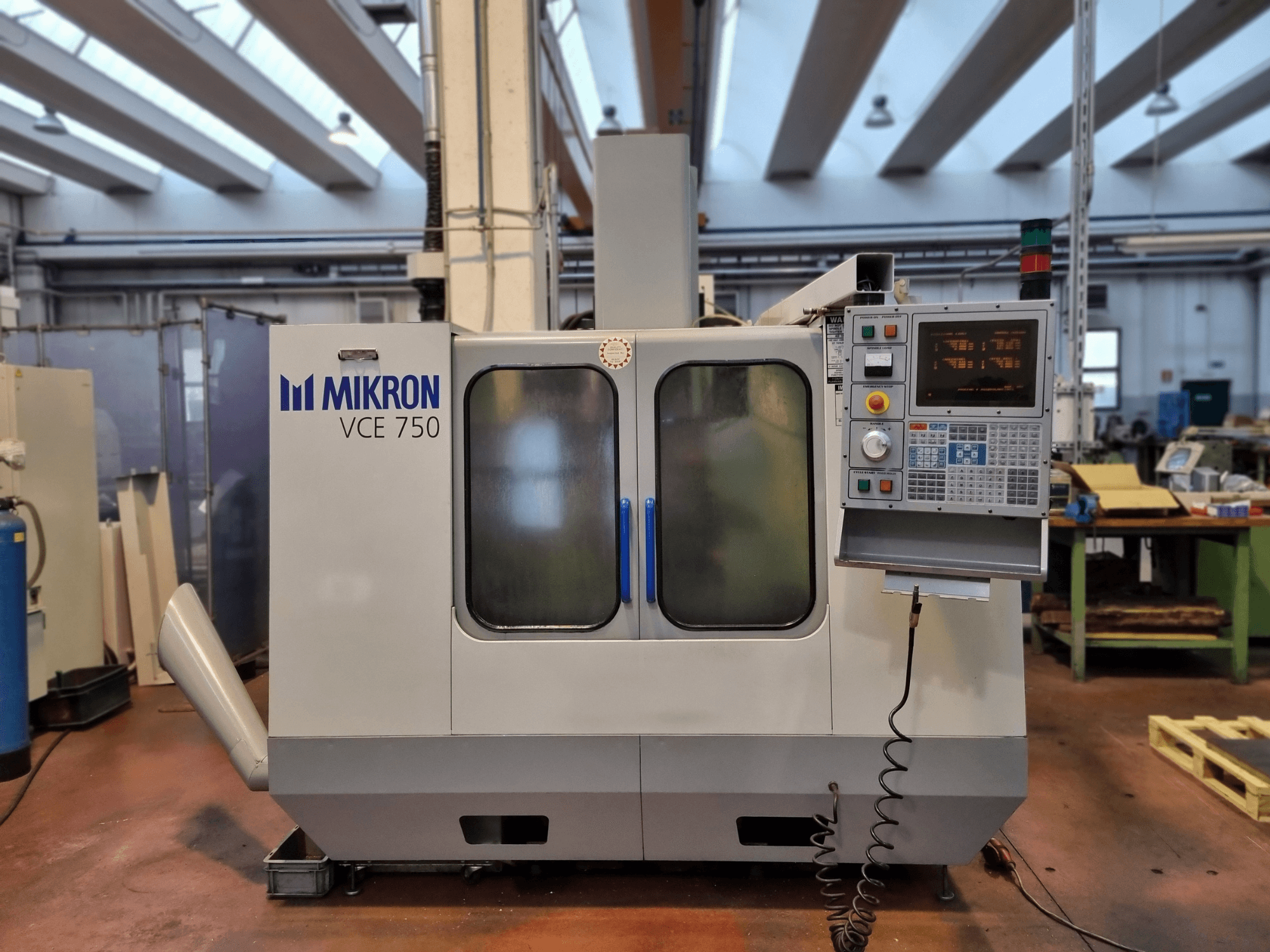 Baltas Mikron VCE 750 CNC darbnīcā, skats no sāniem, ar digitālo vadības paneli un lielām stikla durvīm.