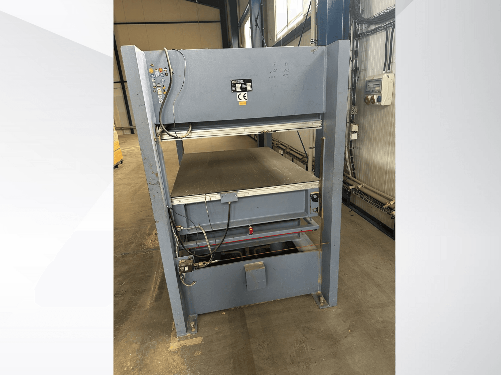 Pelēka rūpnieciska mašīna ar vadības paneli, skatoties no priekšpuses; ietver regulējamas platformas un drošības marķējumu.