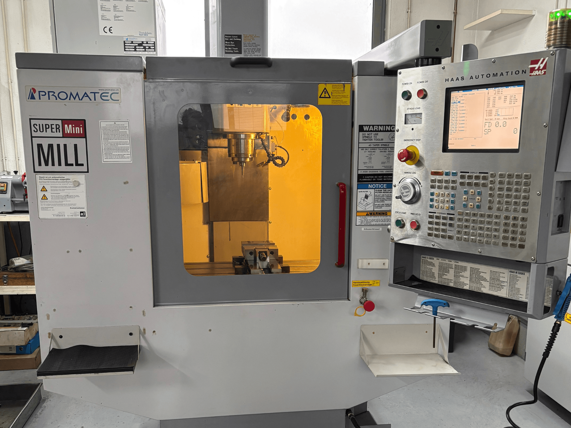 Promatec Super Mini frēze priekšpusē, aprīkota ar Haas Automation vadības paneli un redzamu griezējinstrumentu iekšpusē.