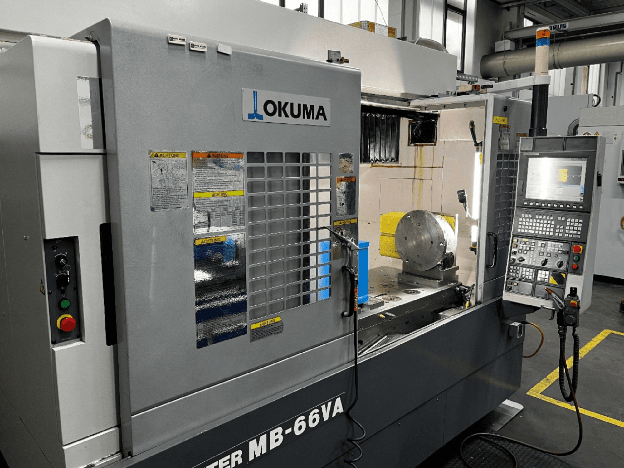 Okuma MB66-VA vertikālās apstrādes centra priekšējais skats, kurā redzams vadības panelis, apstrādājamā detaļa un mašīnas iekšpuse ar drošības uzlīmēm.