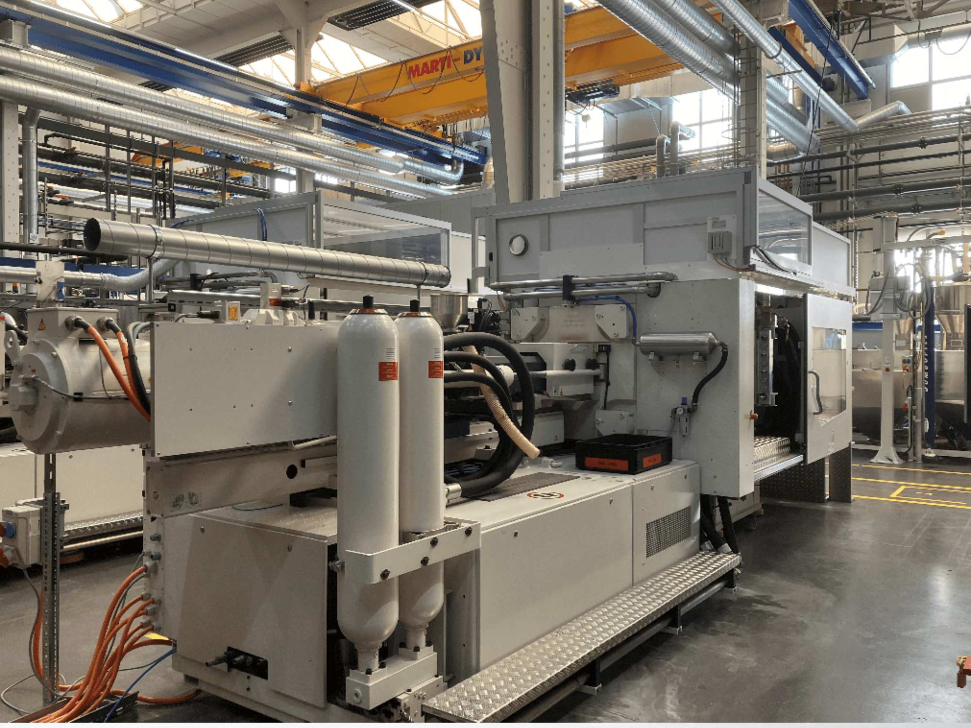 Rūpnieciskā mašīna sānskatā ar vairākām šļūtenēm, divām vertikālām tvertnēm un vadības paneli; zīmols/modelis nav redzams.