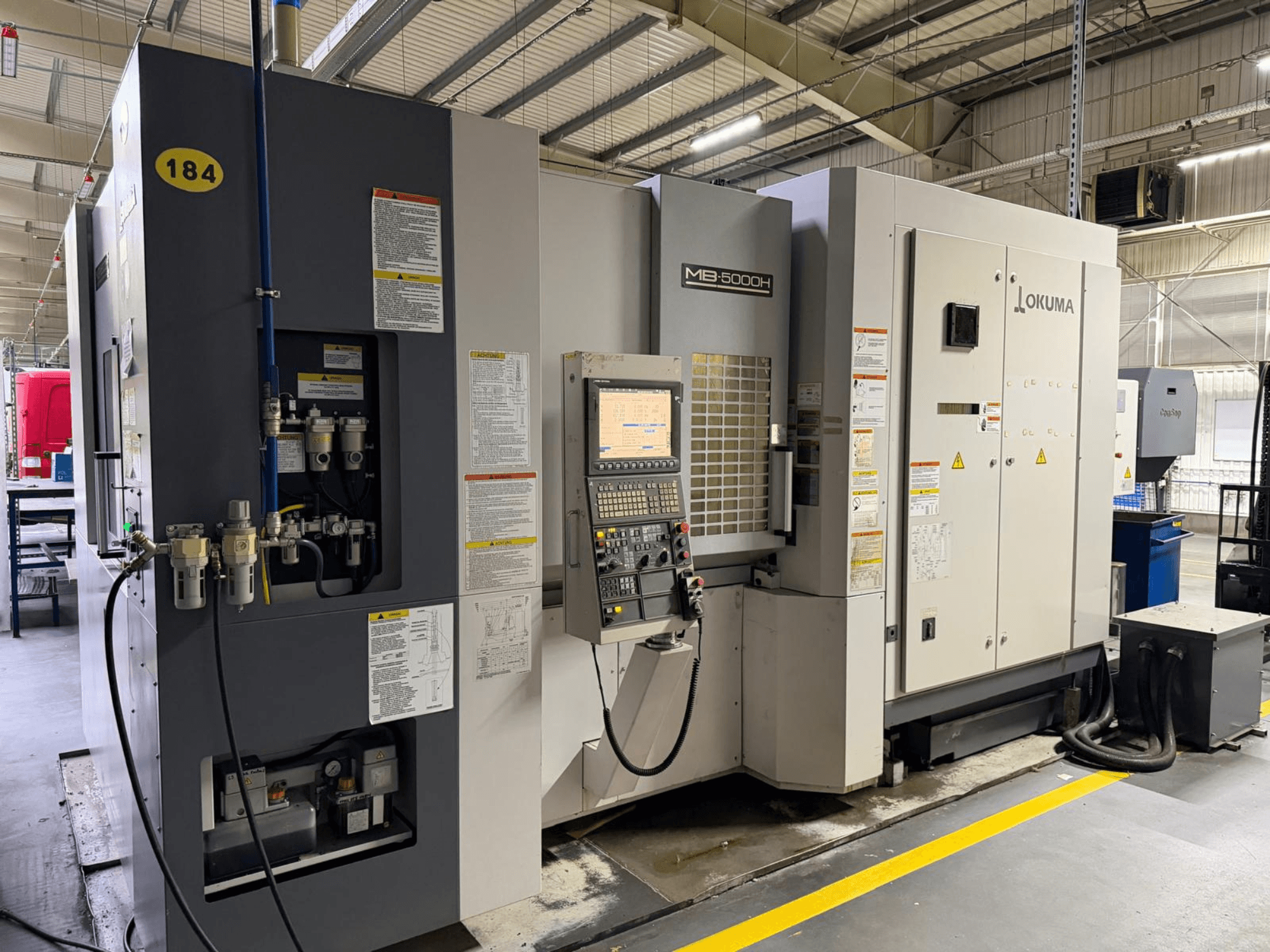 Okuma MB 5000H horizontālā apstrādes centra priekšējais skats ar vadības ierīcēm, marķējumu un hidrauliskajiem savienojumiem.