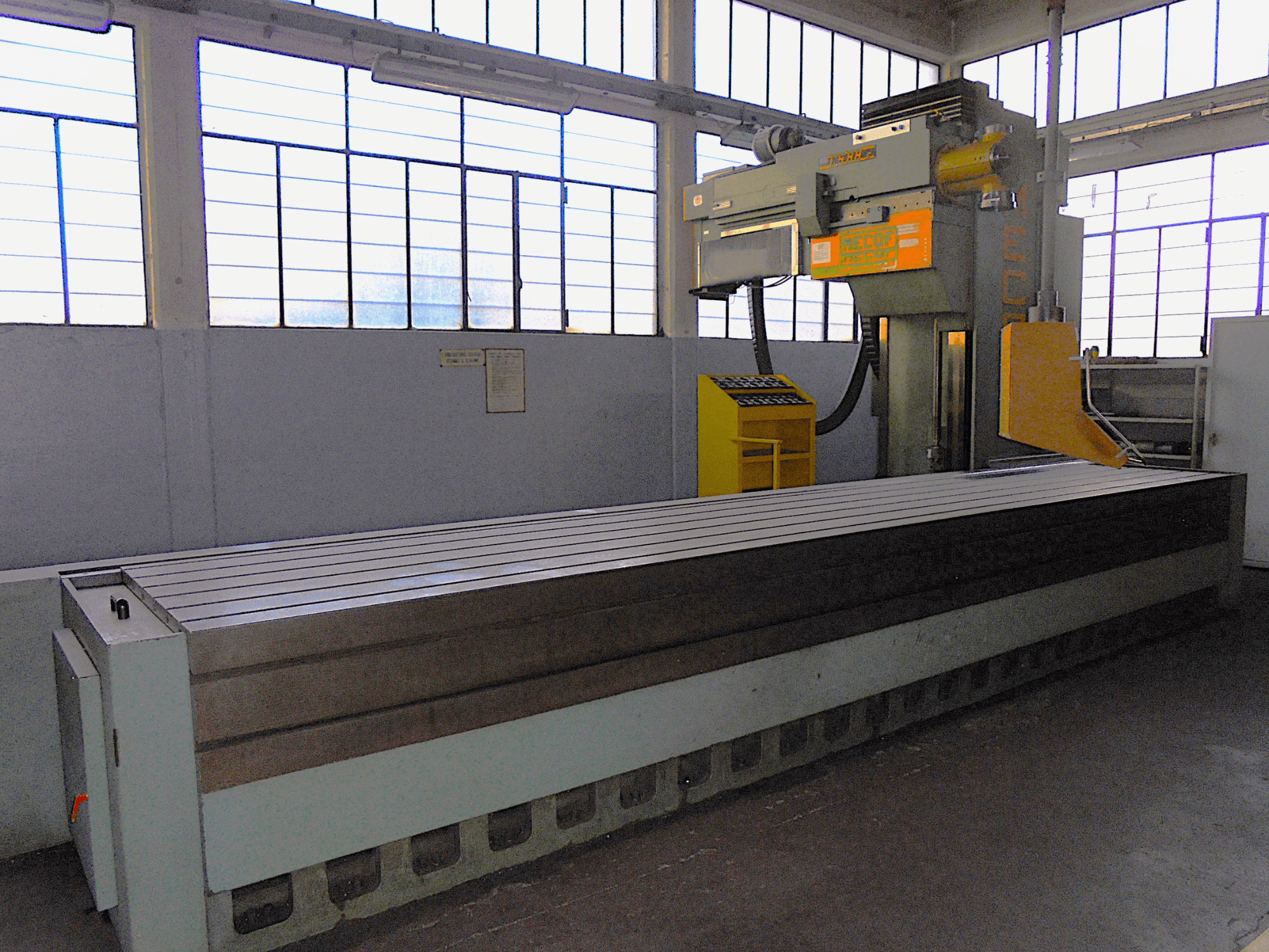 Nezināma zīmola vertikālā frēzmašīna rūpnīcā, skats no sāniem, ar lielu galdu ar rievotu virsmu un vadības paneli.