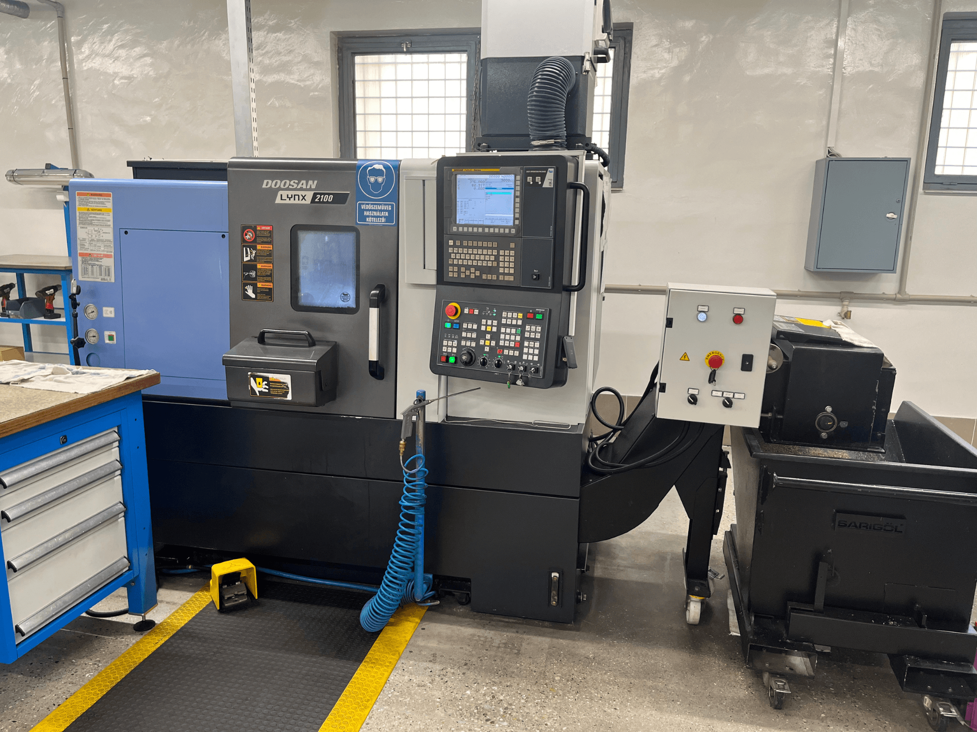 Doosan Lynx 210 virpa darbnīcā, redzams vadības panelis, instrumentu glabāšanas vieta un dzesēšanas sistēma, sānu skats ar darba vietas detaļām.