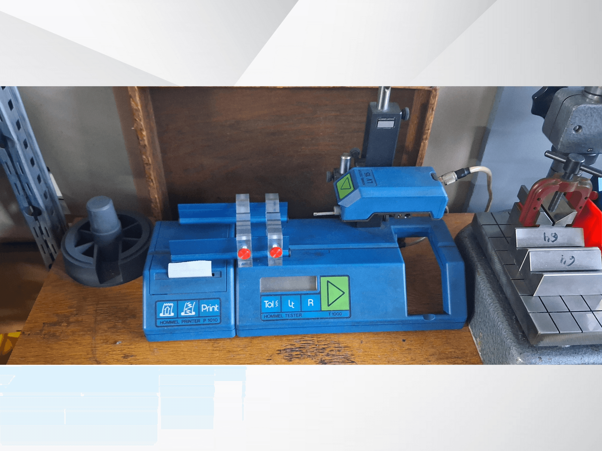 Zils Hommel T 1000 printeris un T 1 010 testeris ar sarkanām pogām, skatīts no priekšpuses uz darba galda ar metāla blokiem.