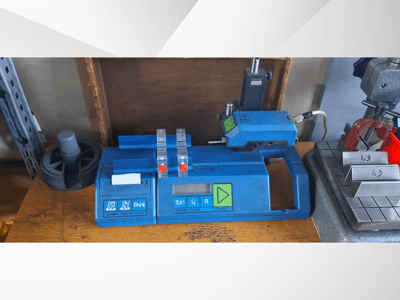 Zils Hommel T 1000 printeris un T 1 010 testeris ar sarkanām pogām, skatīts no priekšpuses uz darba galda ar metāla blokiem.