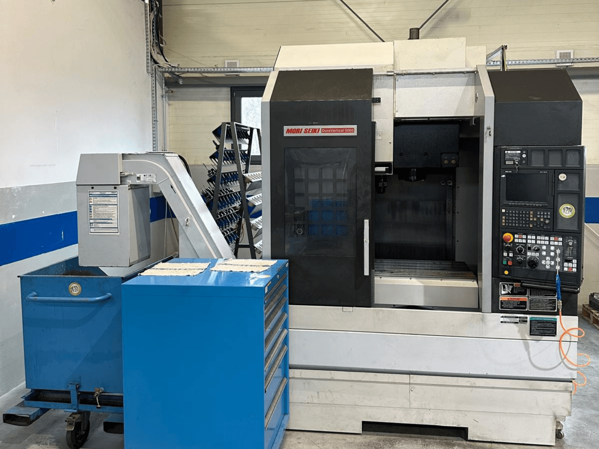 MORI SEIKI DURA VERTICAL 5060 — vertikālā apstrādes centra skats no priekšpuses ar vadības pulti un blakus esošo instrumentu glabāšanas vietu.