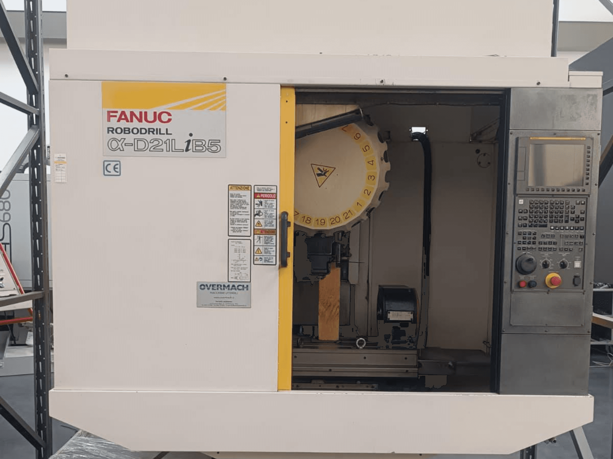 FANUC Robodrill α-D21LiB5, skats no priekšpuses; CNC iekārta ar redzamu instrumentu turētāju, vadības paneli un drošības brīdinājumiem.