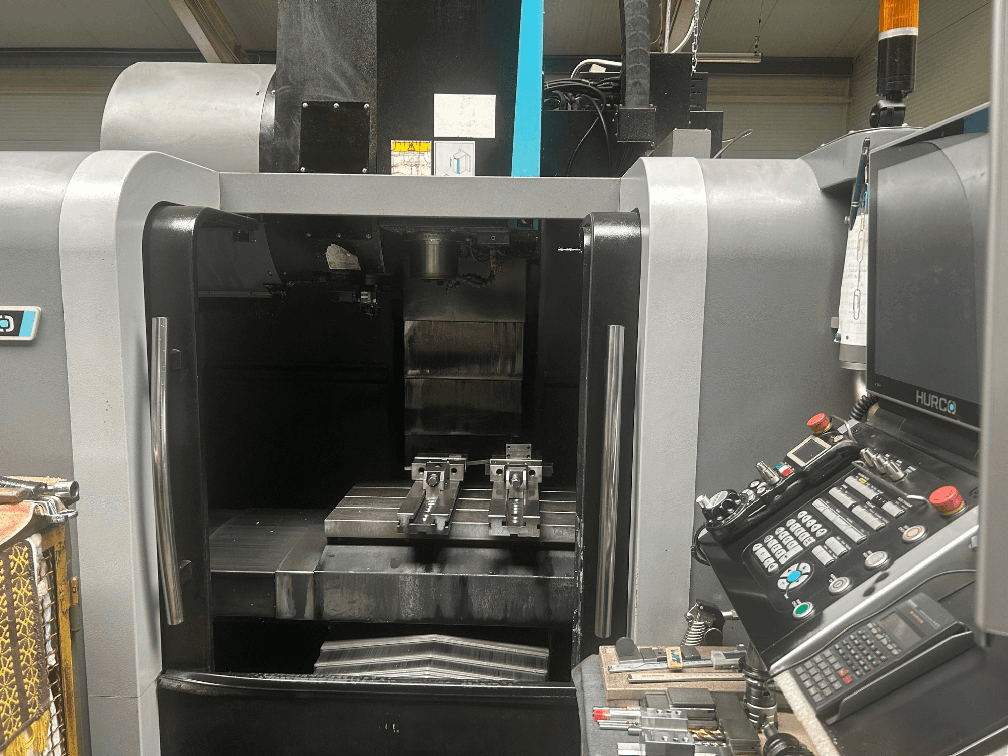 Hurco CNC apstrādes centra priekšējais skats, kurā redzams instrumentu turētājs, metāla sagataves un vadības panelis ar pogām un displeju.