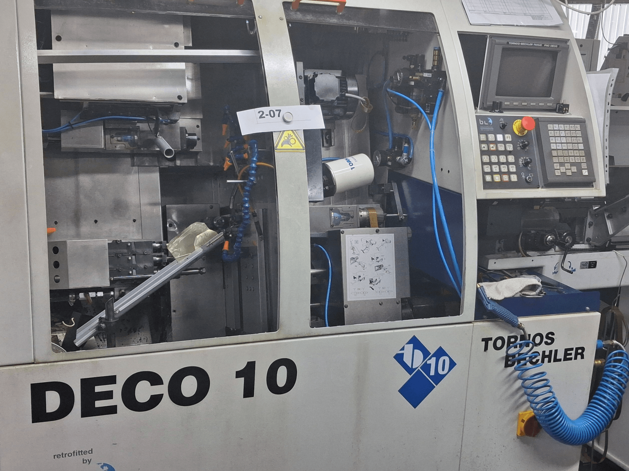 Tornos Deco 10a Šveices tipa virpošanas mašīnas TORNOS Deco 10a tuvplāna skats, kurā redzams iekšējais mehānisms un vadības paneļa detaļas.