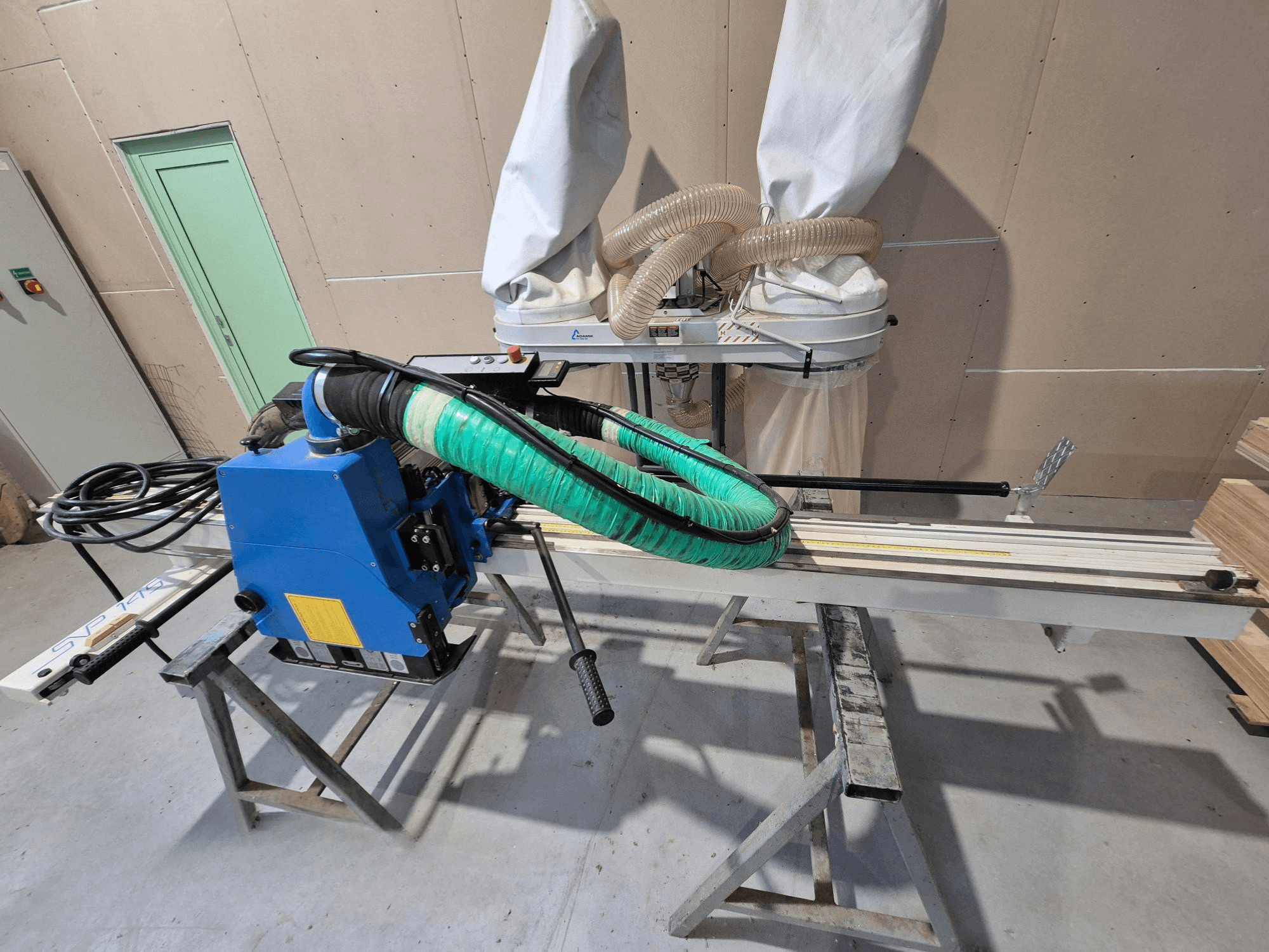 Zils SVP-175 horizontālais paneļu zāģis ar zaļu putekļu šļūteni un balstu, skatīts no sāniem darbnīcā.