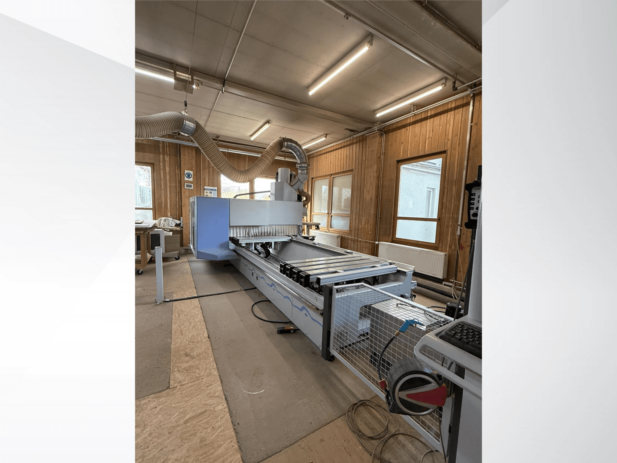 CNC frēzmašīna darbnīcā, skats no sāniem, ar lielu zilu korpusu, pievienotu lentes konveijeru un redzamu vadības paneli.