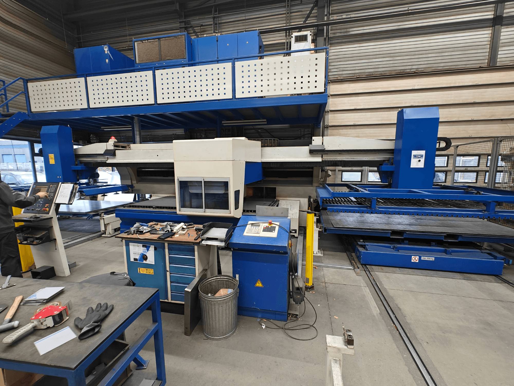 Rūpnieciskās CNC darbgaldi no priekšpuses, ar zili baltu konstrukciju, vadības paneli un dažādiem instrumentiem uz darba galda.