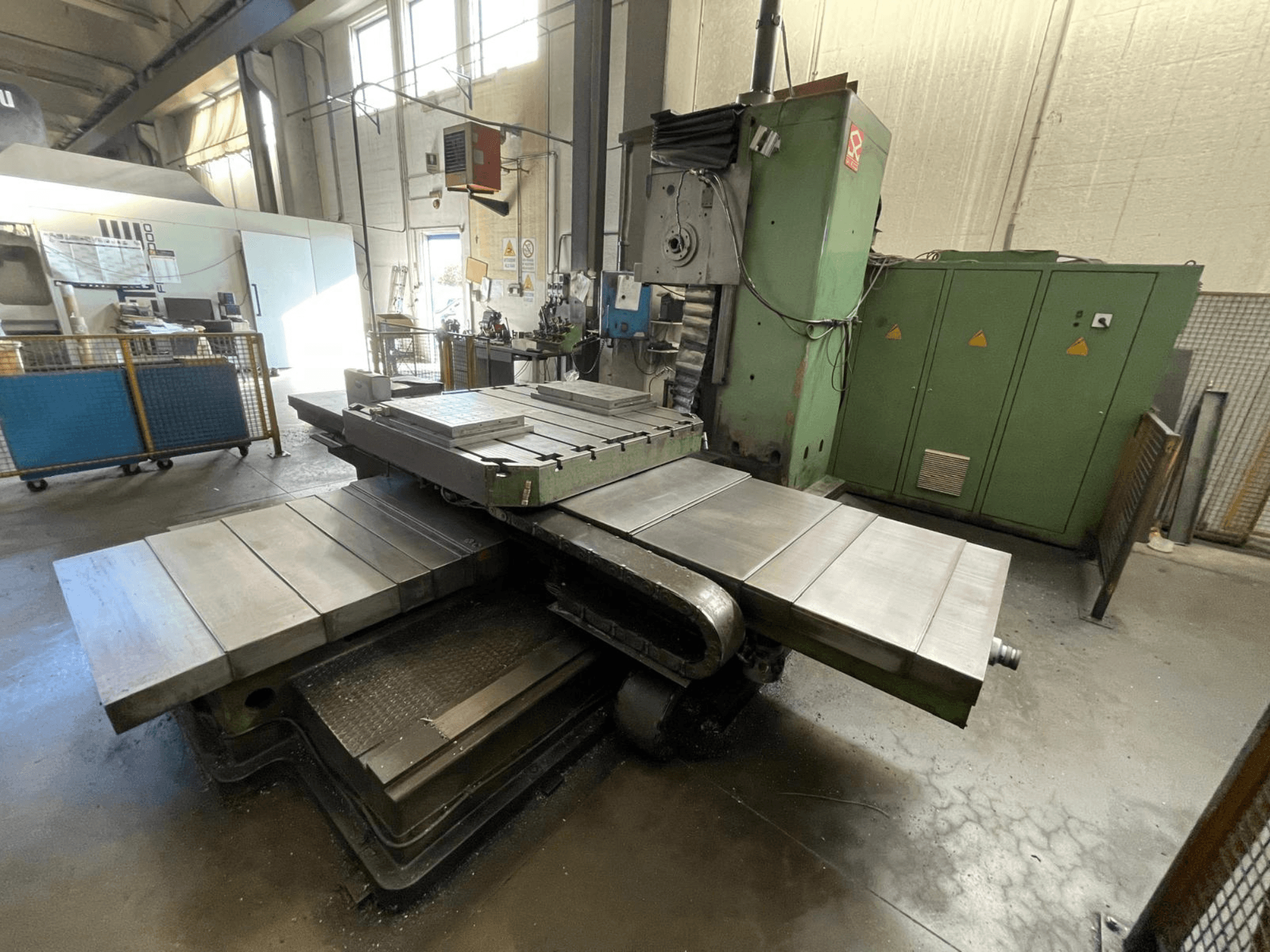 Liela rūpnieciska frēzmašīna ar zaļu korpusu, skats no sāniem, ar vairākām plakanām virsmām un glabāšanas skapjiem fonā.