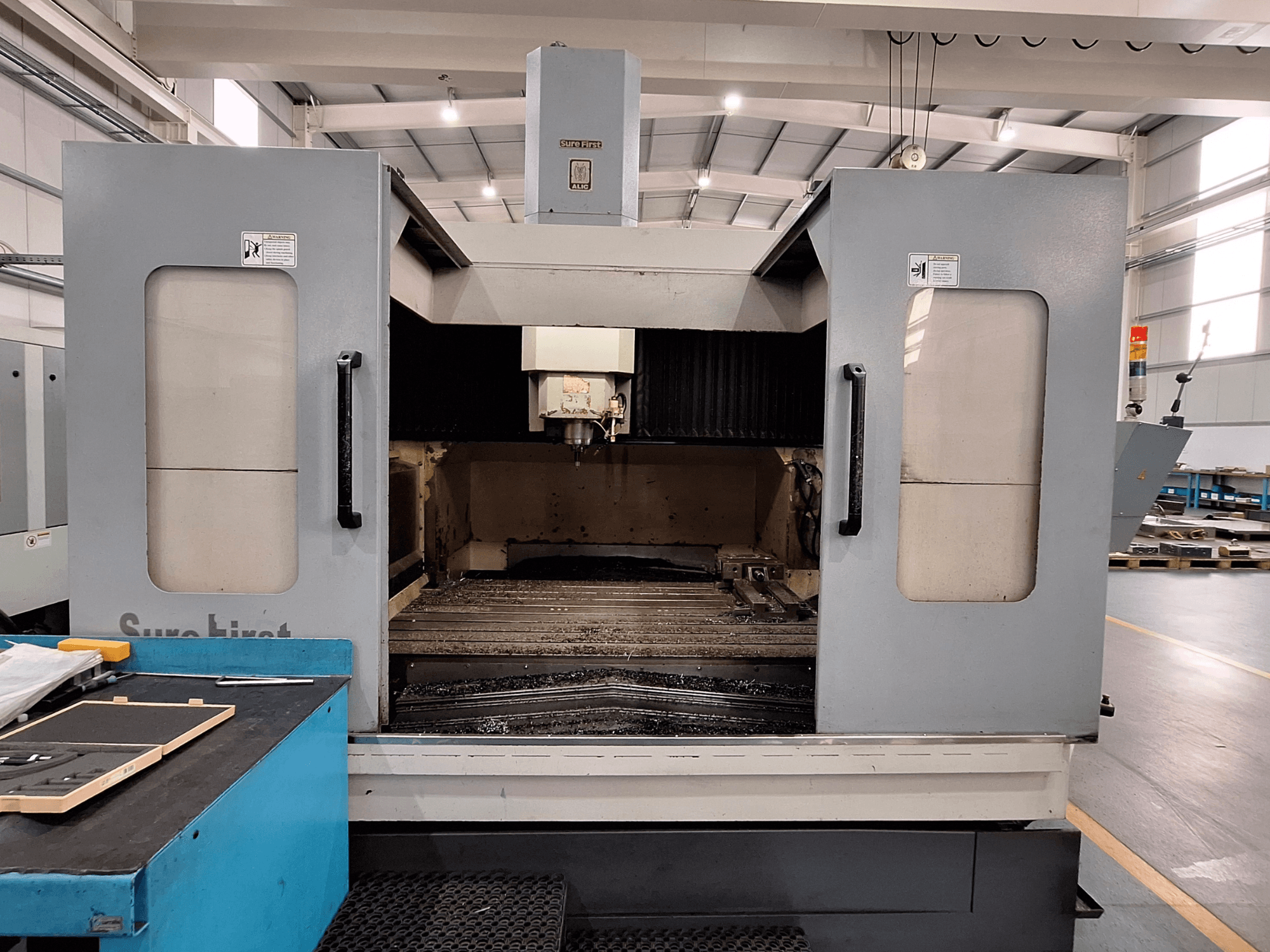 Vertikālais apstrādes centrs Sure First QM-1836, priekšējais skats, kurā redzama instrumentu galva un tukša darba zona, mūsdienīga rūpnieciska vide.