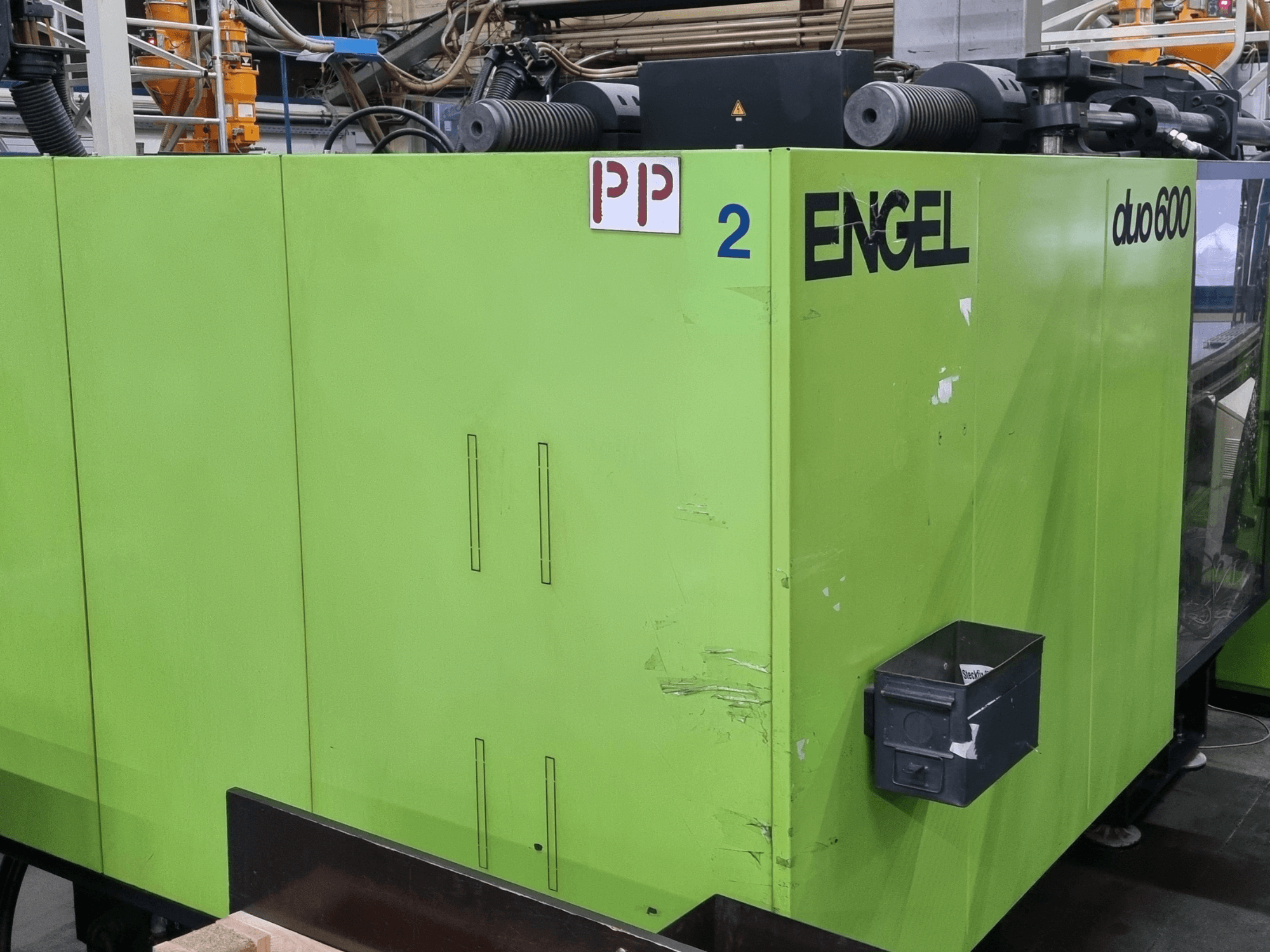 Hidrauliskā plastmasas liešanas mašīna „Engel duo 600“, zaļa ārējā apdare, skats no priekšpuses, ar marķētām detaļām un materiālu uzglabāšanas kasti.