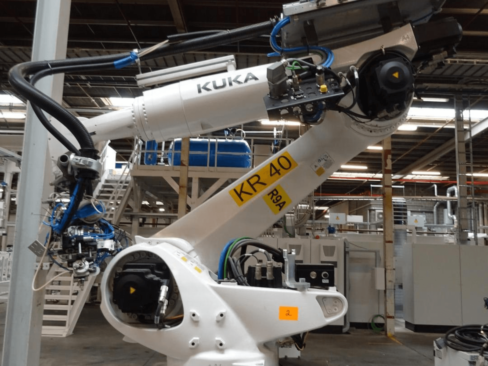 KUKA KR 120 R3100-2 robota roka sānu skatā, ar vadības paneli un mehāniskiem komponentiem, uzstādīta industriālā vidē.