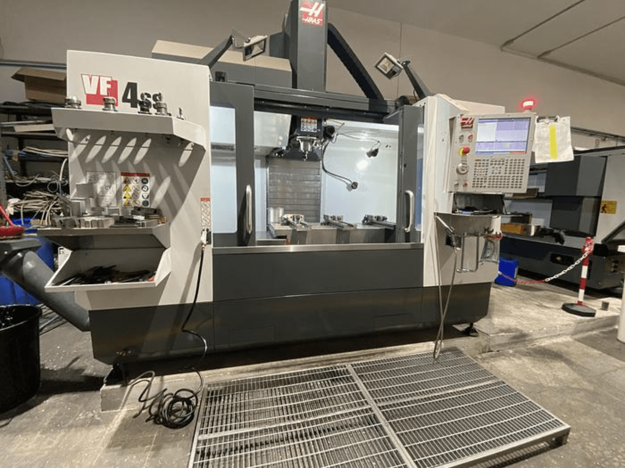 Haas VF-4SS CNC frēzmašīna, skats no priekšpuses, ar instrumentu glabāšanas vietu, vadības paneli un redzamu darba zonu darbnīcā.