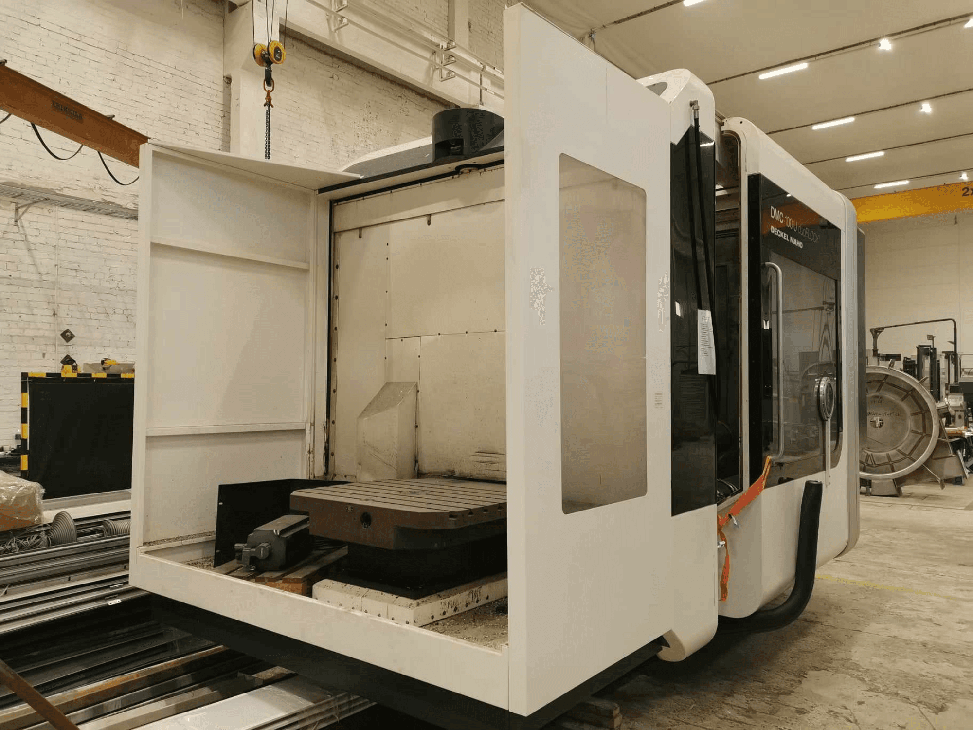 DMC 100U lineārās CNC darbgaldi, skats no sāniem ar atvērtām piekļuves durvīm, redzams darba galds un iekšējās sastāvdaļas.