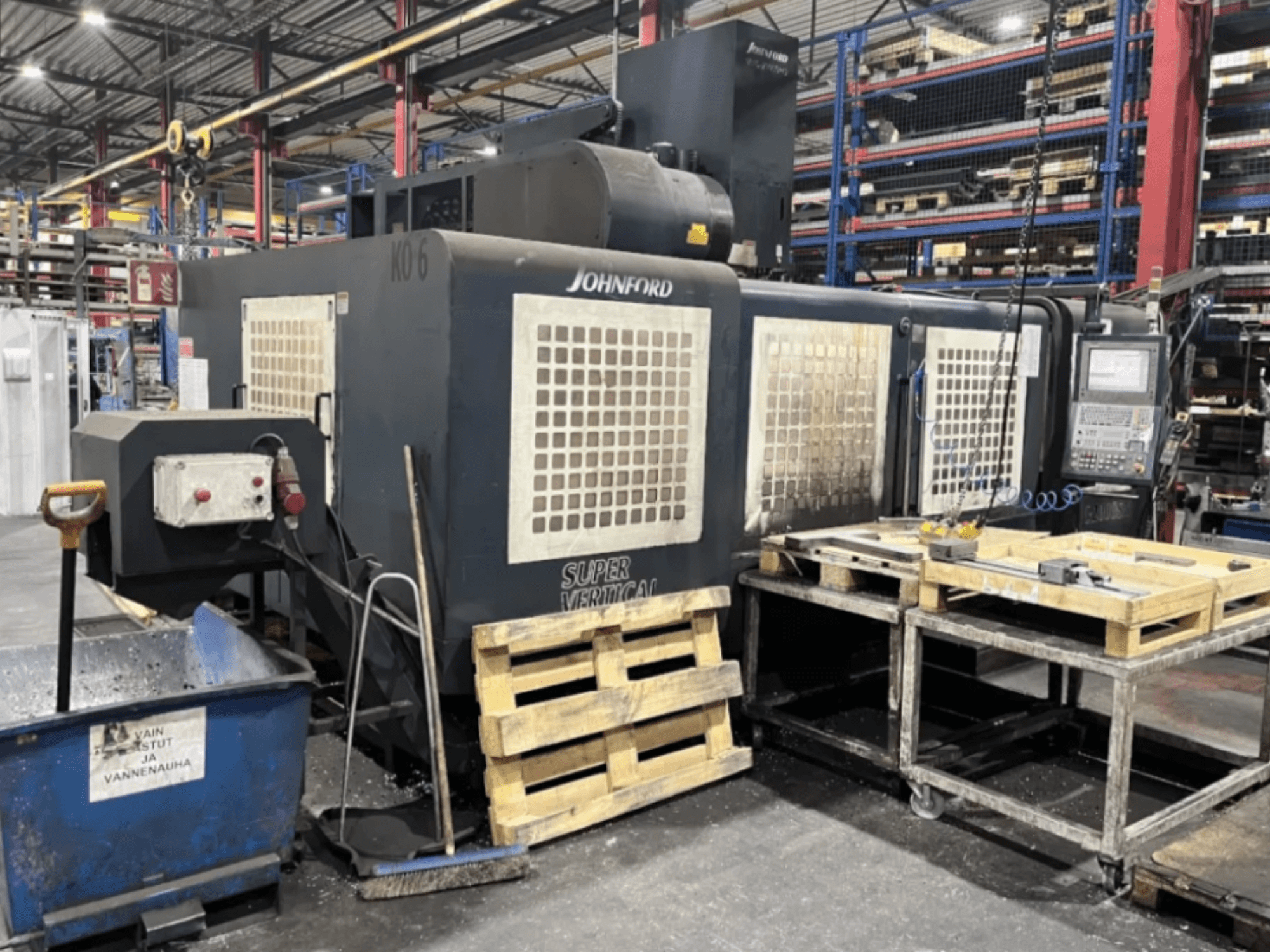 Johnford Super Vertikālā frēzmašīna rūpnīcā, skats no sāniem ar vadības paneli, paliktņiem un tīrīšanas iekārtām blakus.