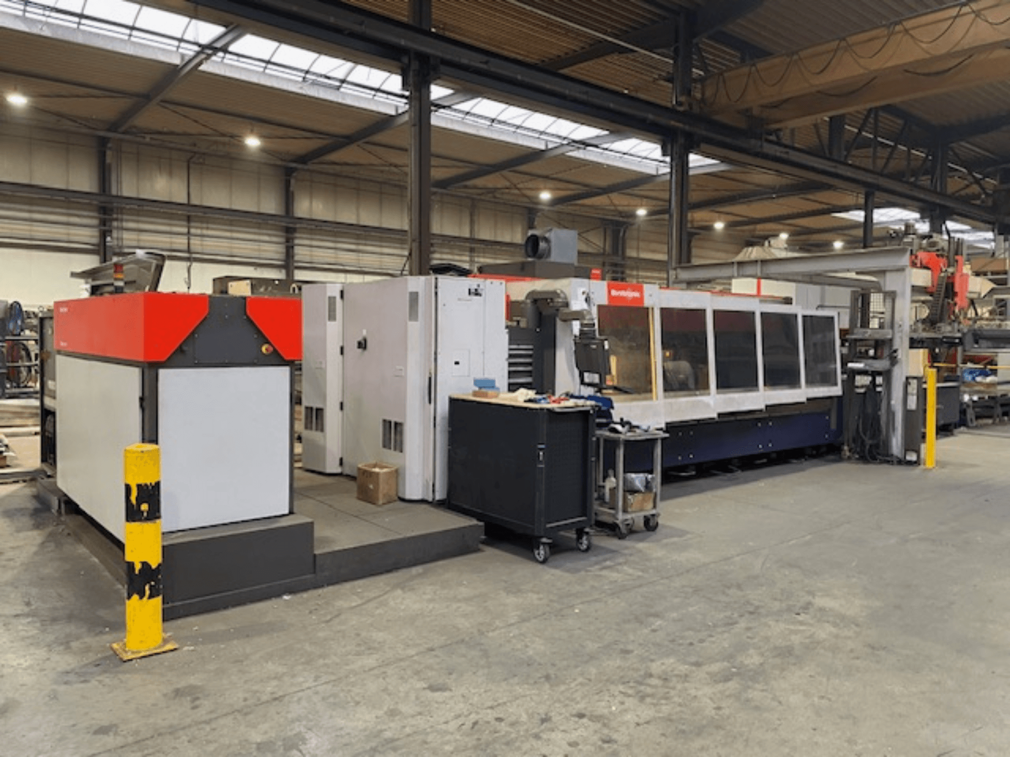 Bystronic BySpeed 3015 CO2 lāzera griešanas iekārta, skats no sāniem darbnīcā, ar izteiktiem sarkanā un pelēkā dizaina elementiem.