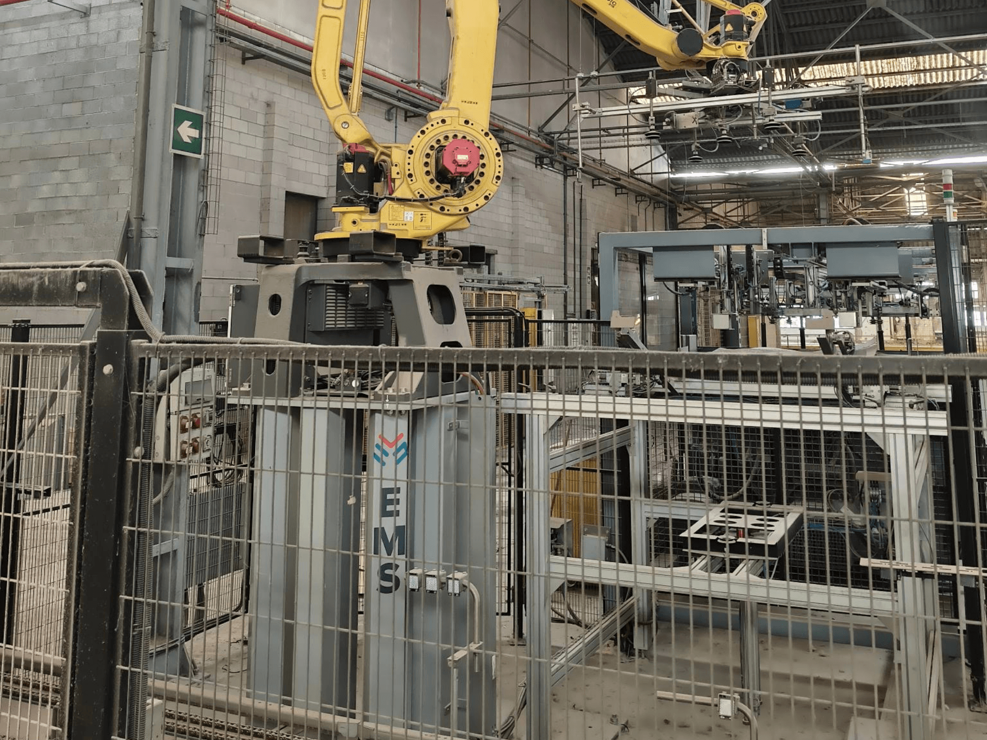 FANUC M-410iC/315 robota rokas priekšskats, kas novietota virs EMS instrumenta, fonā redzamas rūpnieciskās iekārtas un nožogojums.
