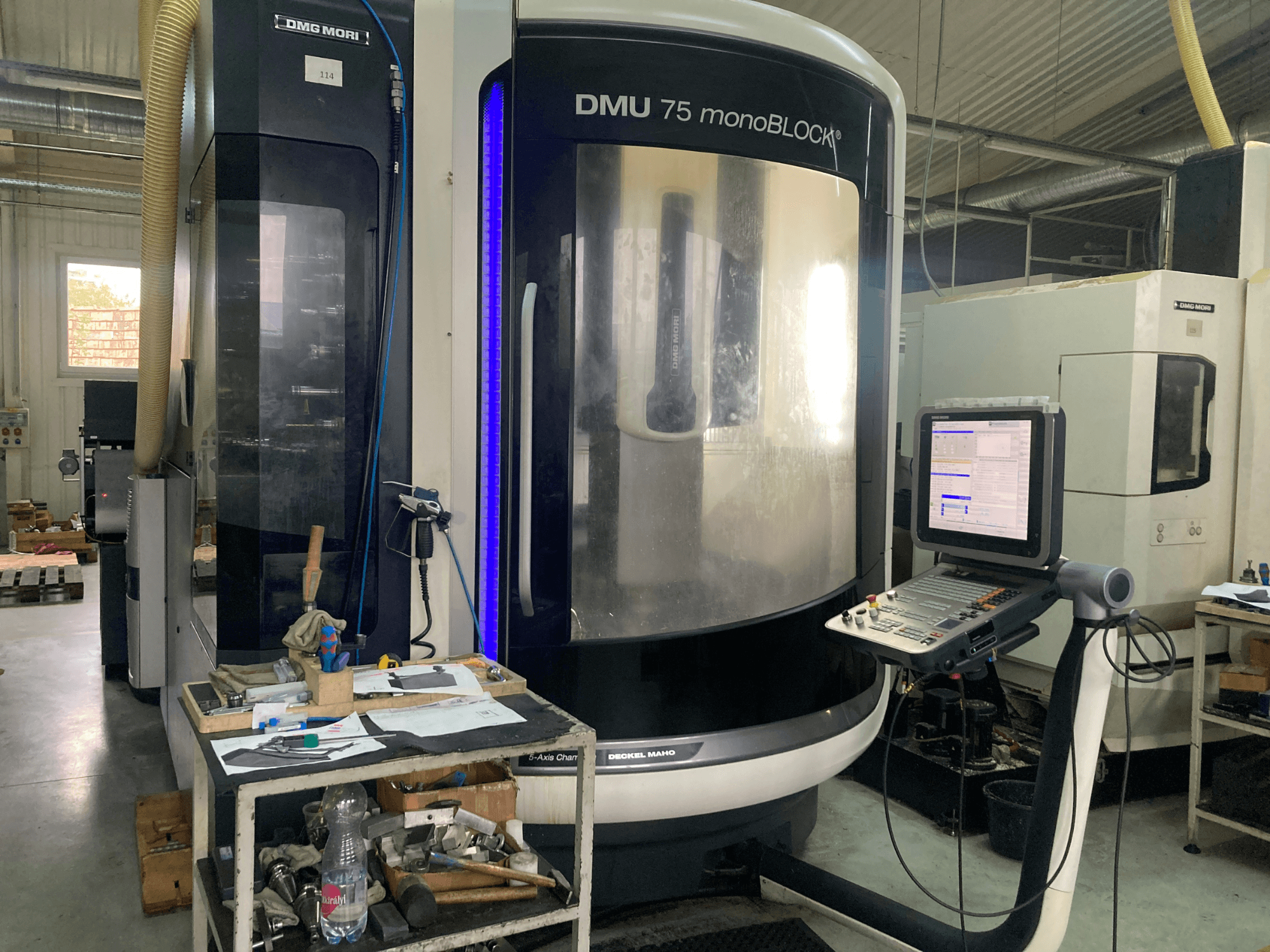 DMG MORI DMU 75 monoBLOCK® CNC mašīna, skats no priekšpuses, ar vadības paneli, instrumentiem un darba vietu ar materiāliem.