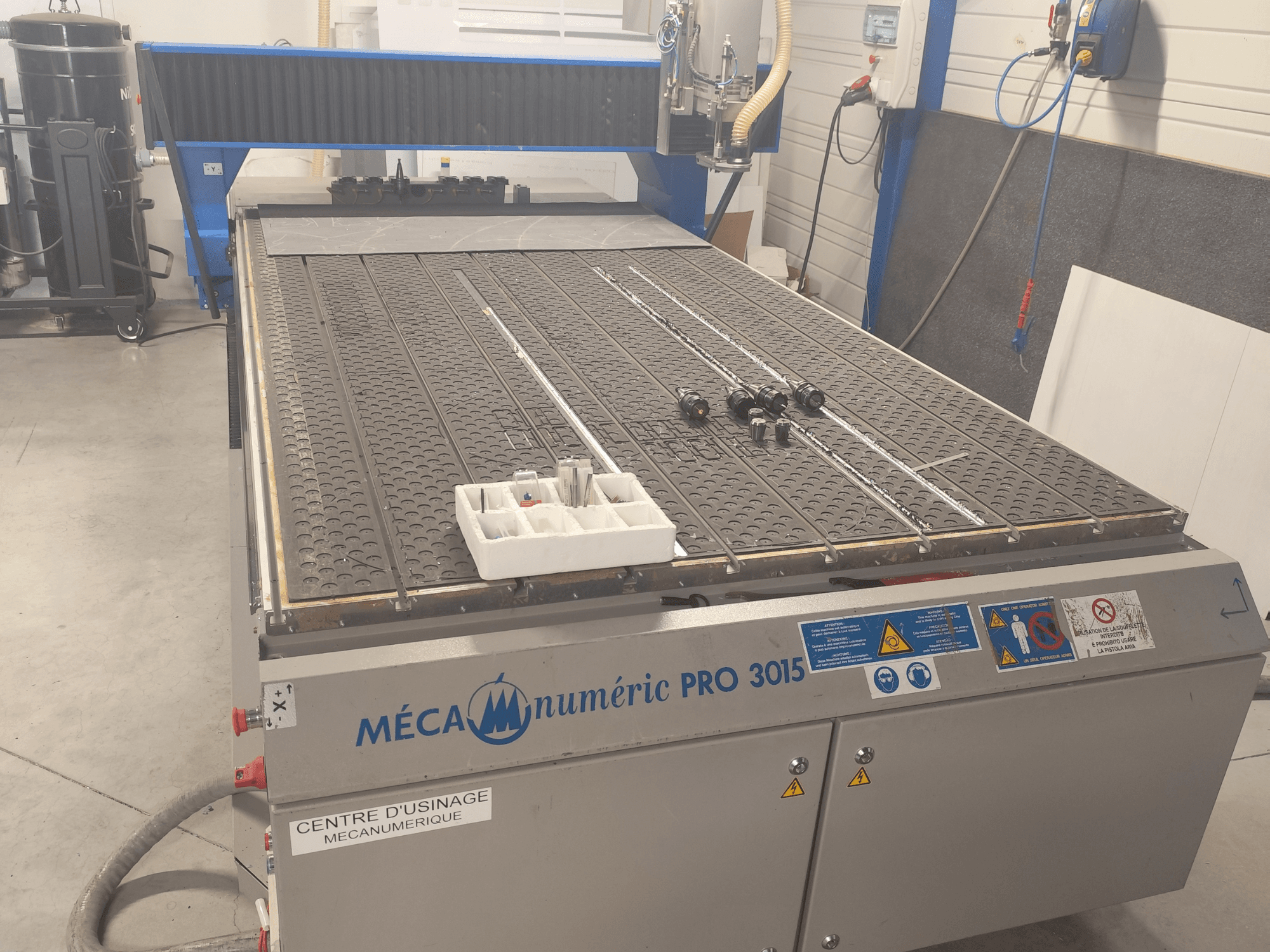 CNC frēzes MECANUMERIC Pro 3015 skats no priekšpuses, kurā redzams detalizēti attēlots griešanas galds, vadības panelis un piederumi uz galda.