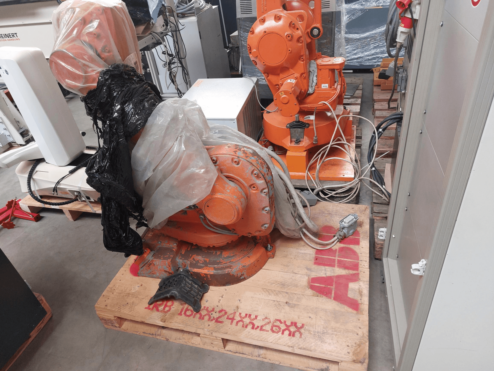 ABB IRB 1600 M2004 robota roka, kas daļēji pārklāta ar plastmasu, skatoties no priekšpuses, ar kabeļiem un papildu iekārtām blakus.