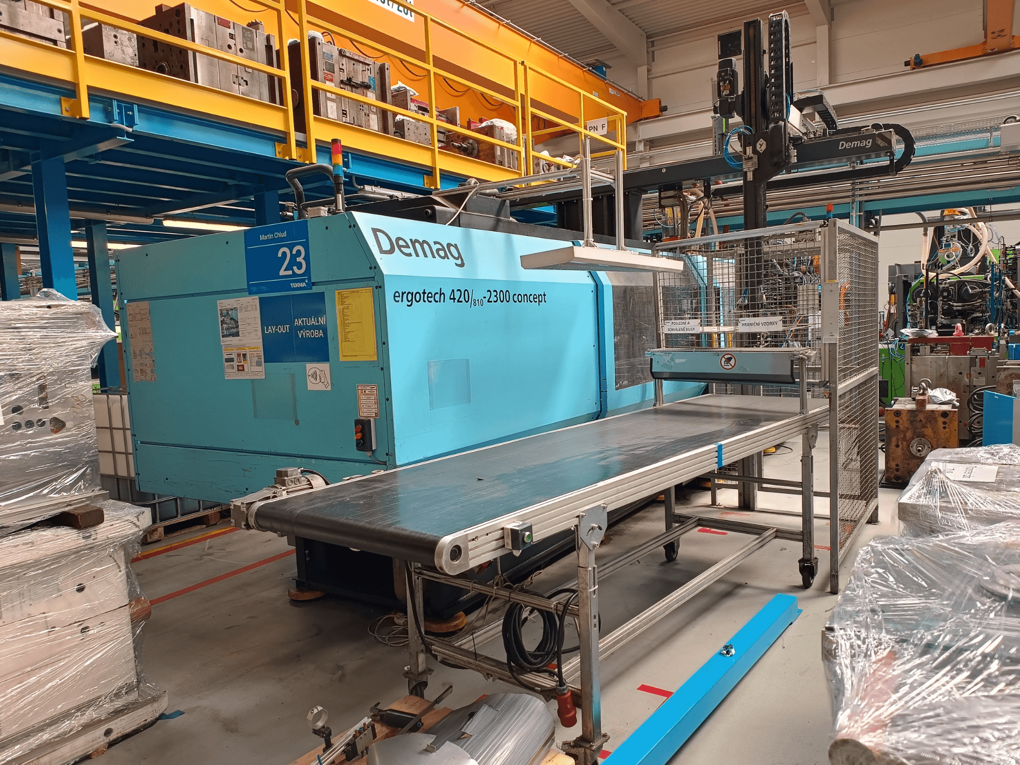 Demag ergotech 420/810 - 2300 hidrauliskā iesmidzināšanas iekārta sānu skatā, ar konveijeru un drošības nodalījumu.