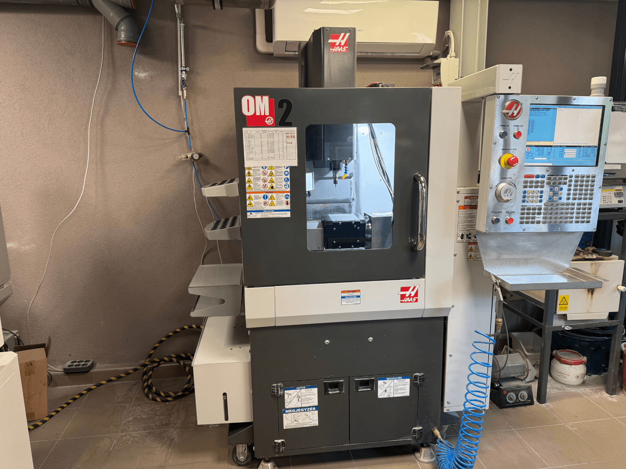 Haas OM-2 CNC darbagalds, skats no priekšpuses, ar vadības paneli, vārpstu un drošības marķējumiem, darbnīcas vidē.