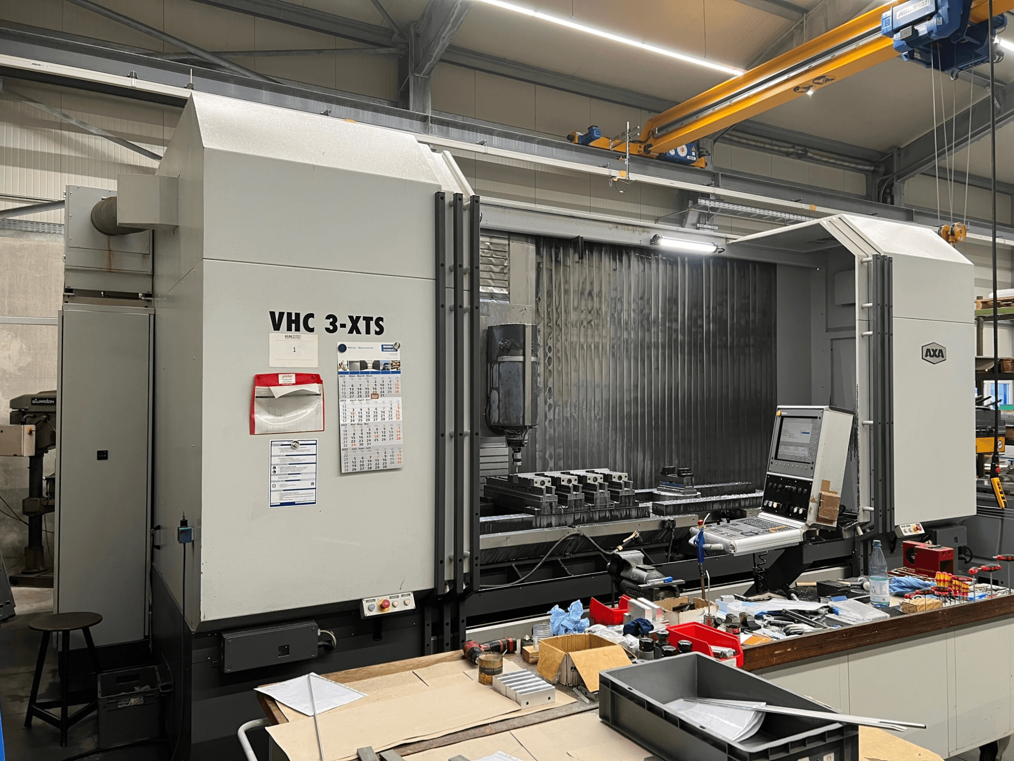 AXA VHC 3-XTS CNC darbagalds AXA VHC 3-XTS, skats no sāniem, ar vadības paneli un darba zonu, kas pārpildīta ar instrumentiem un detaļām.