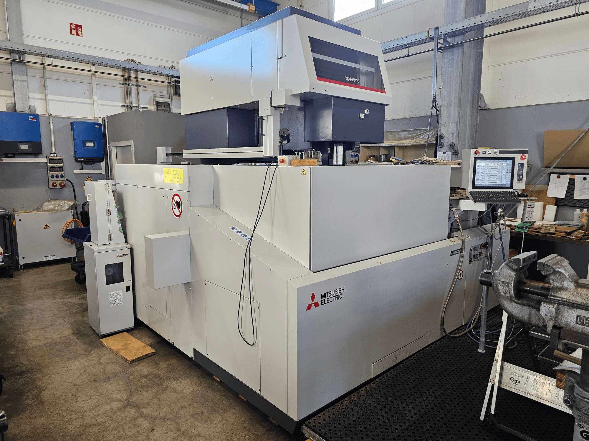 Mitsubishi MV4800 stiepļu EDM mašīna darbnīcā, sānu skats, kurā redzams vadības panelis, drošības marķējumi un apkārtējās iekārtas.