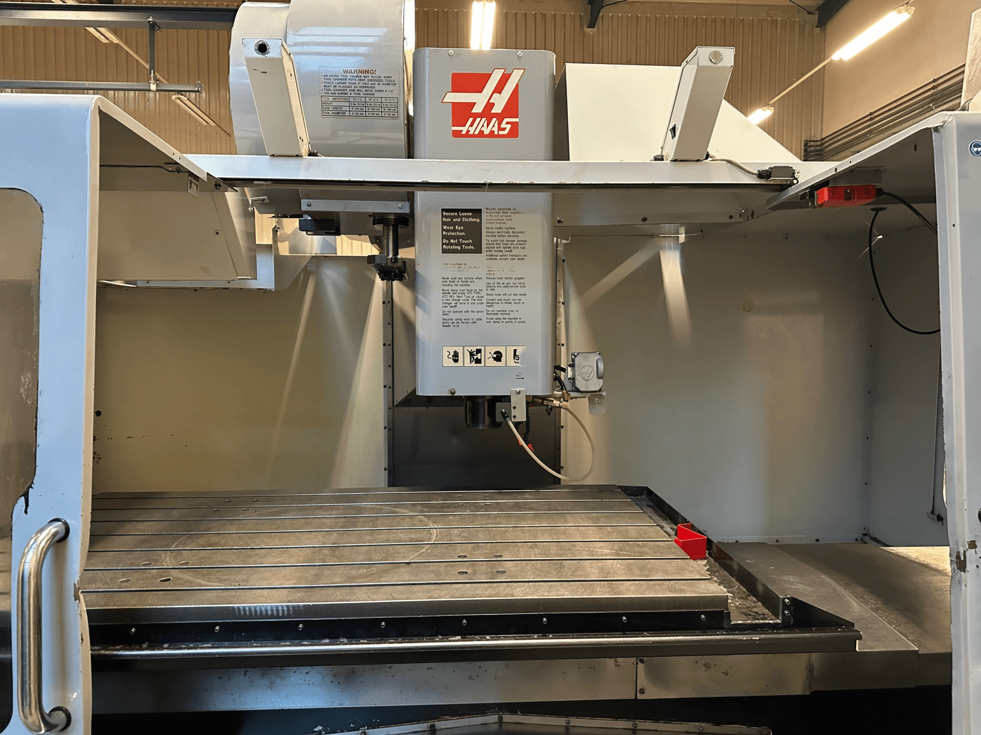 Haas CNC frēzmašīna, modelis VF-2, priekšējais skats, kurā redzamas vadības ierīces, metāla darba galds un drošības marķējums uz paneļa.