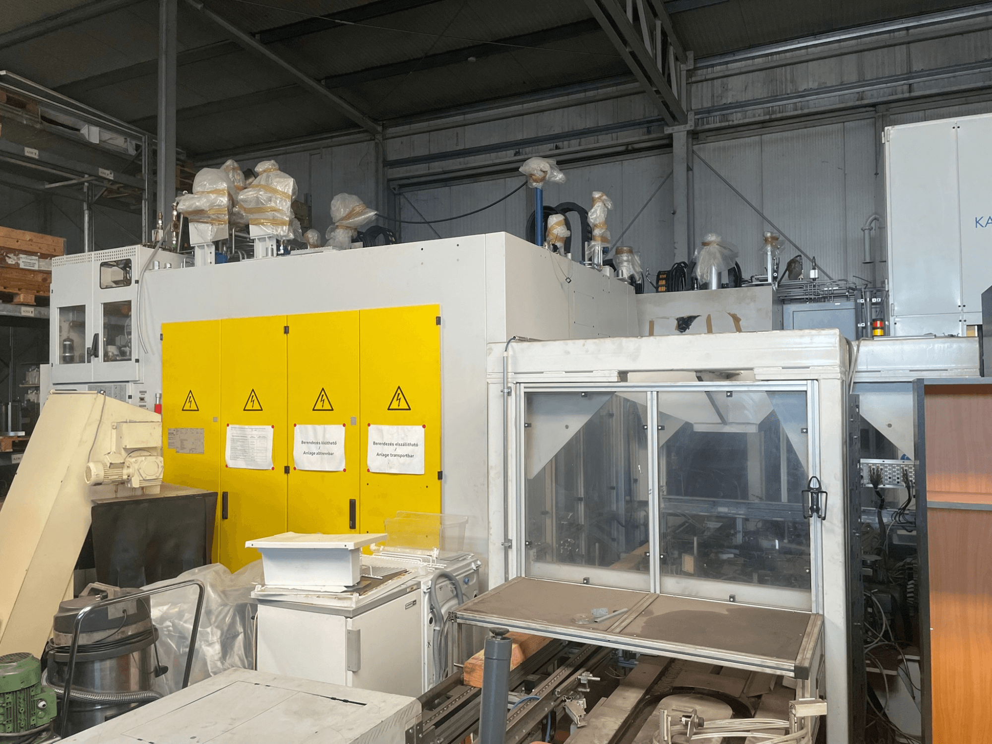 Rūpnieciskās iekārtas ar dzeltenu vadības paneli, skats no sāniem, ar brīdinājuma uzrakstiem un plastmasas pārklājumu, noliktavā.